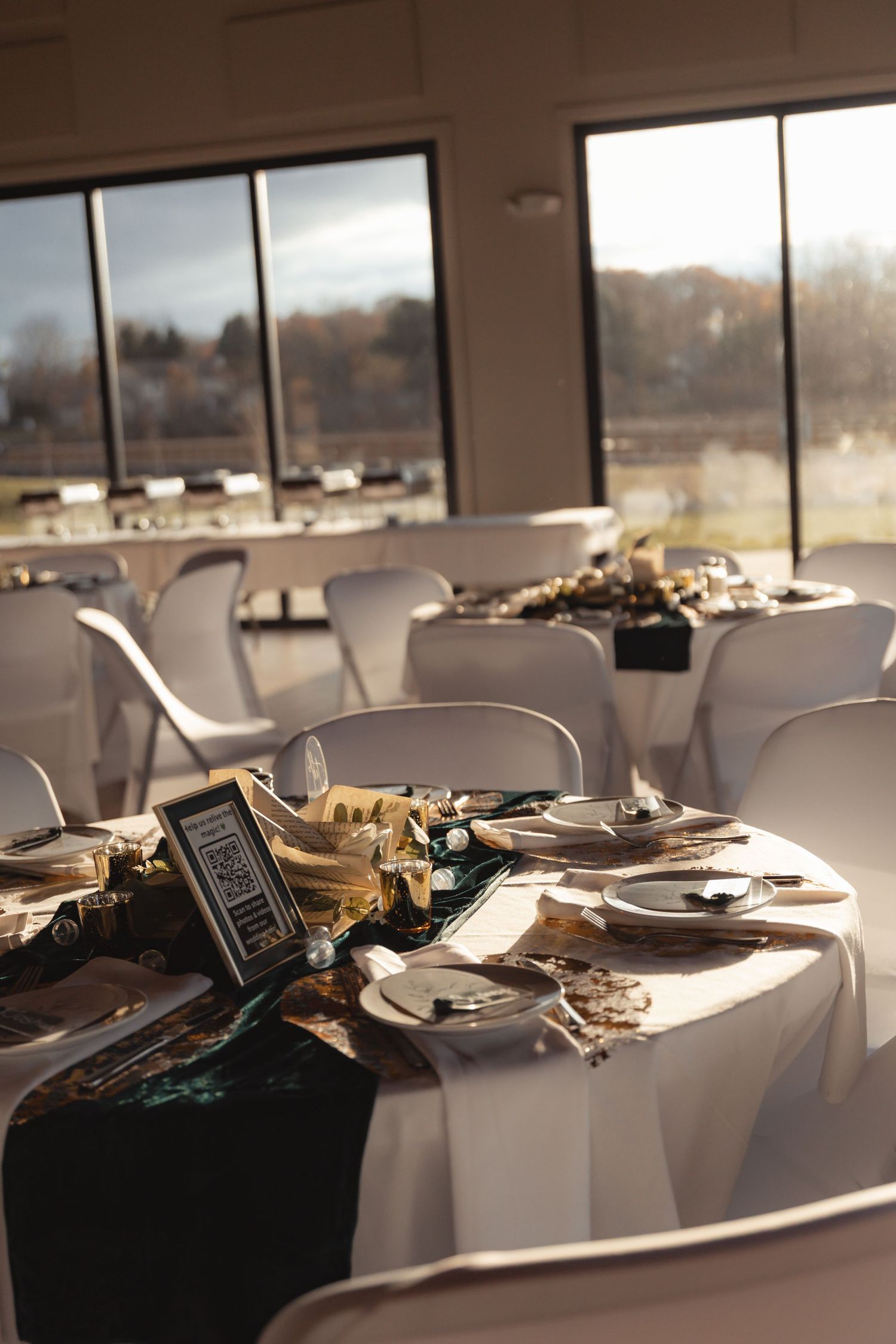 Elegant wedding reception tables with white linens, gold accents, and green runners in a sunlit venue.