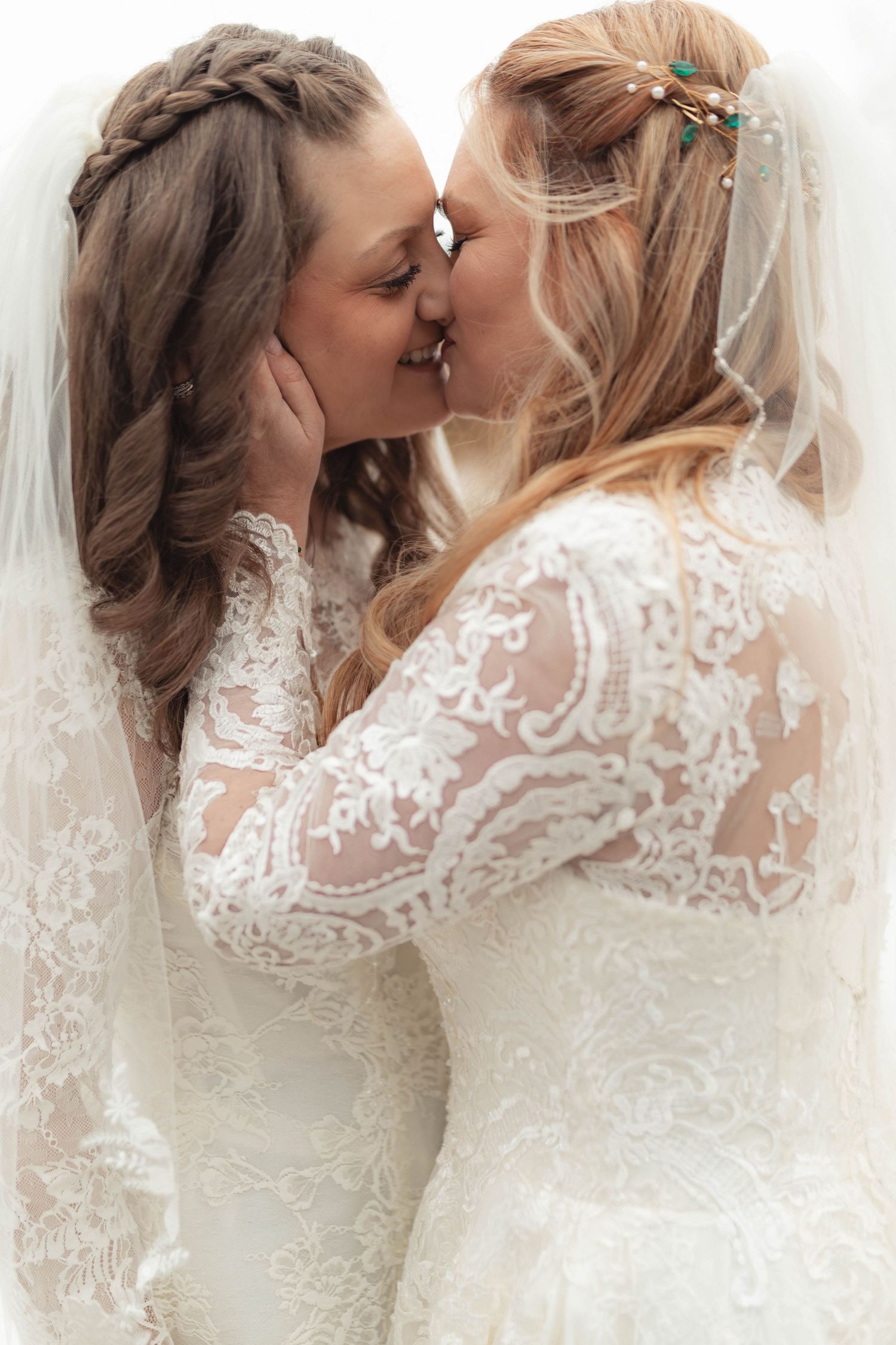 Two brides in lace wedding gowns sharing a romantic kiss, one wearing a veil and floral hair accessories.