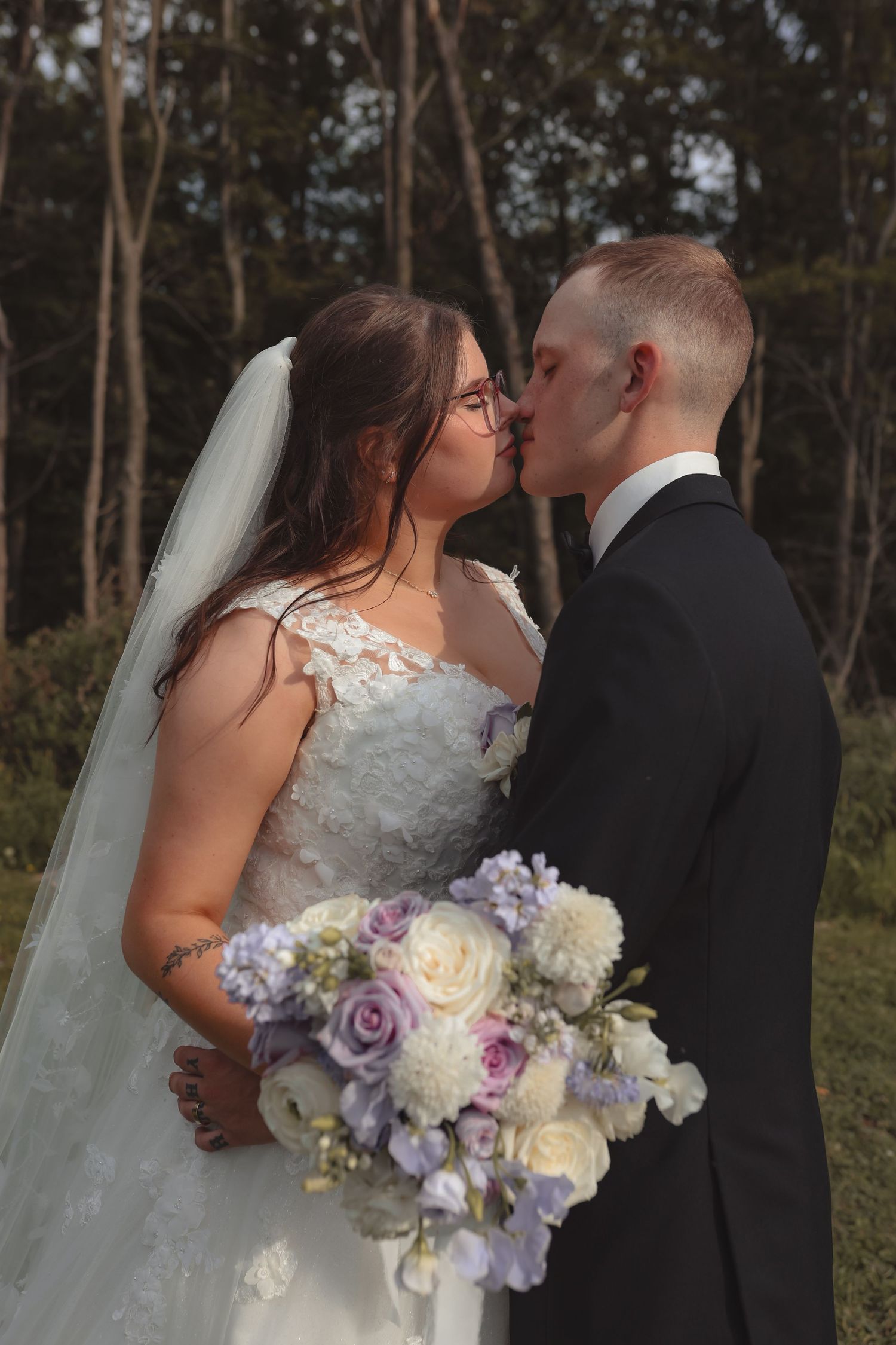 Bride and groom share a kiss outdoors, bride holding a bouquet of white and lavender roses, wearing a lace gown and veil.