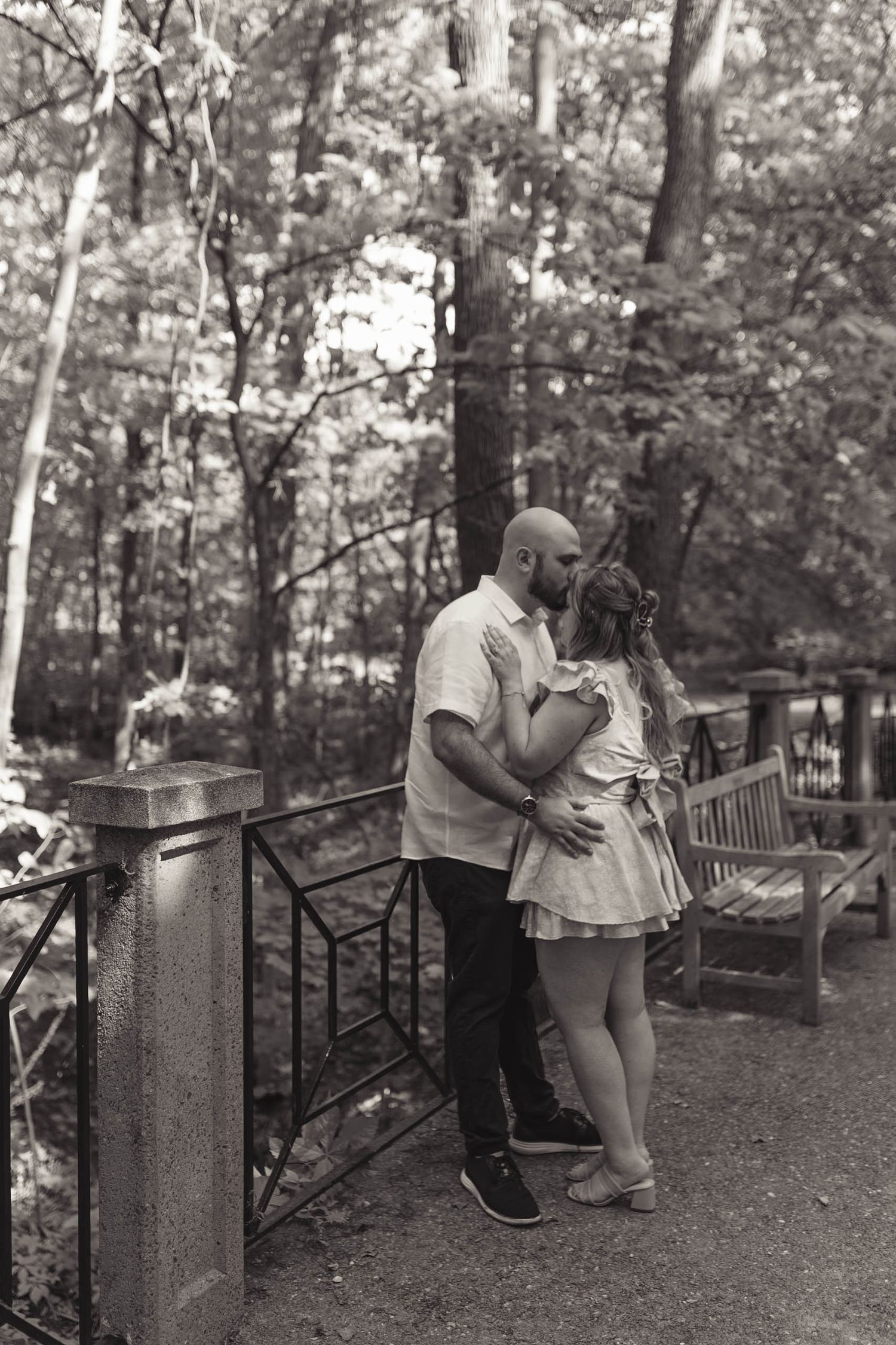 A bald bearded man and a woman in a sundress share a romantic kiss on a park path lined with benches.