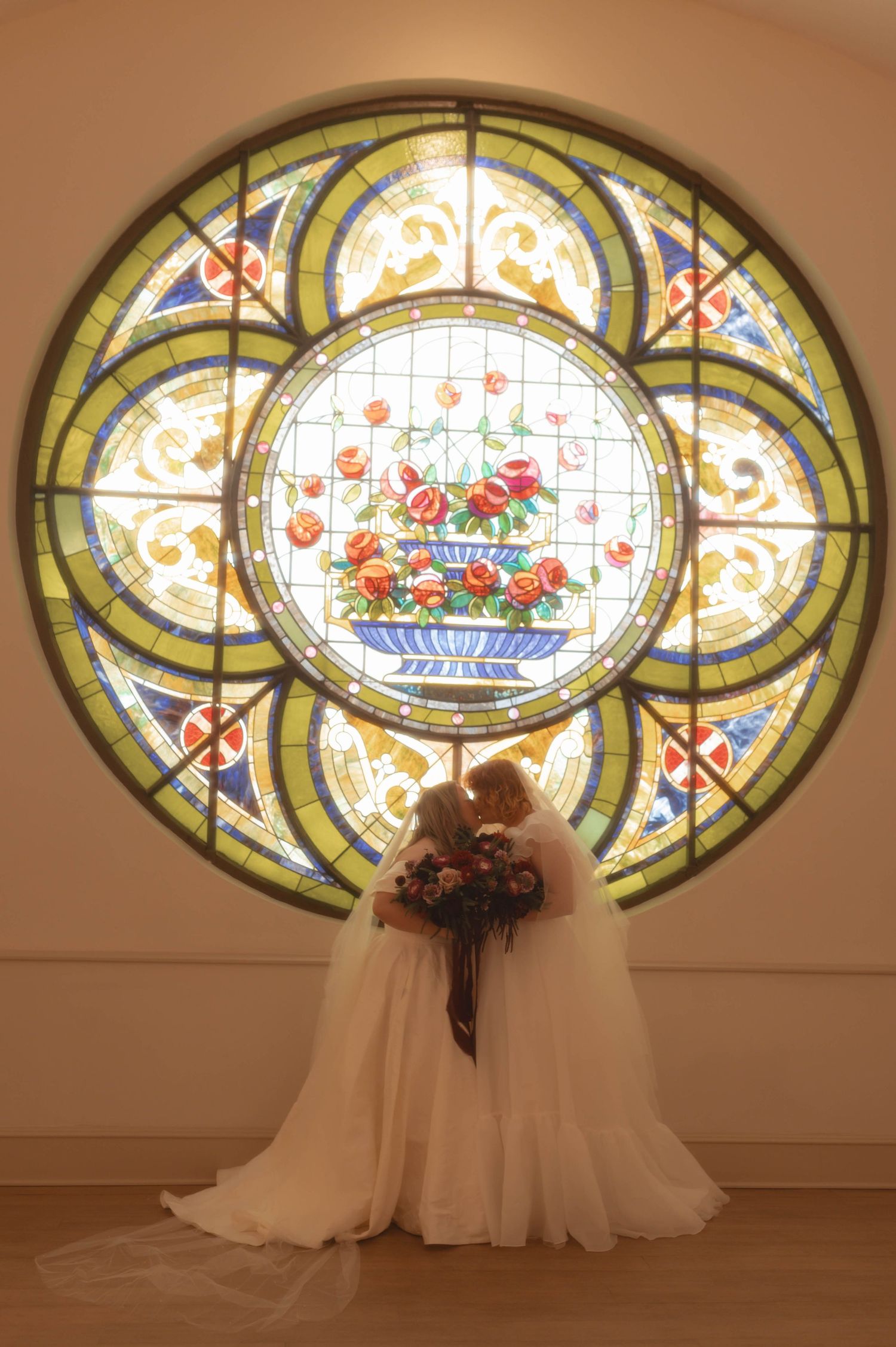 Two brides in white gowns kiss before a stunning circular stained glass window featuring floral motifs in a church.
