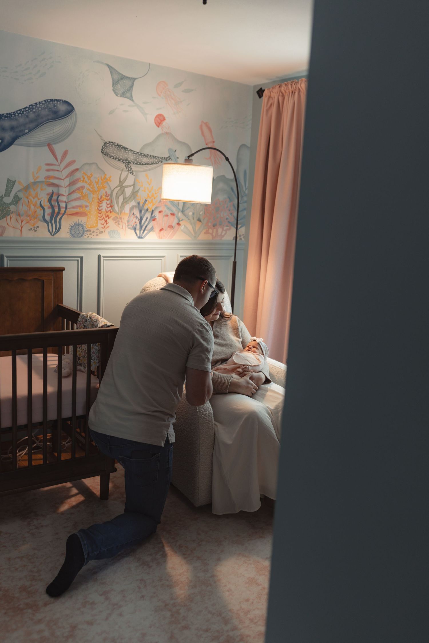 Parents share a tender moment with newborn in a beautifully decorated nursery featuring ocean-themed wallpaper.