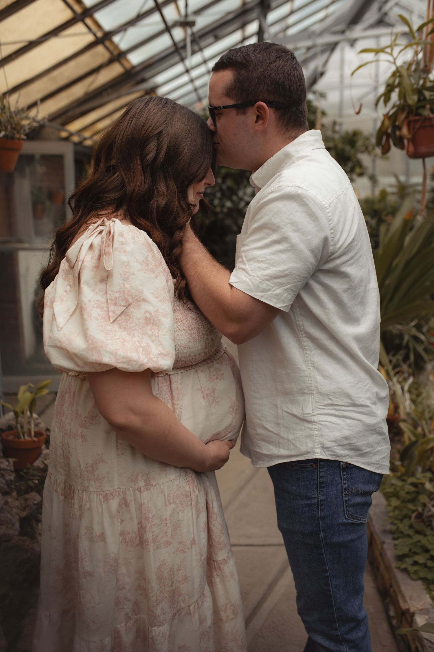 Pregnant woman in floral dress with partner sharing tender moment in lush greenhouse during maternity photo session.
