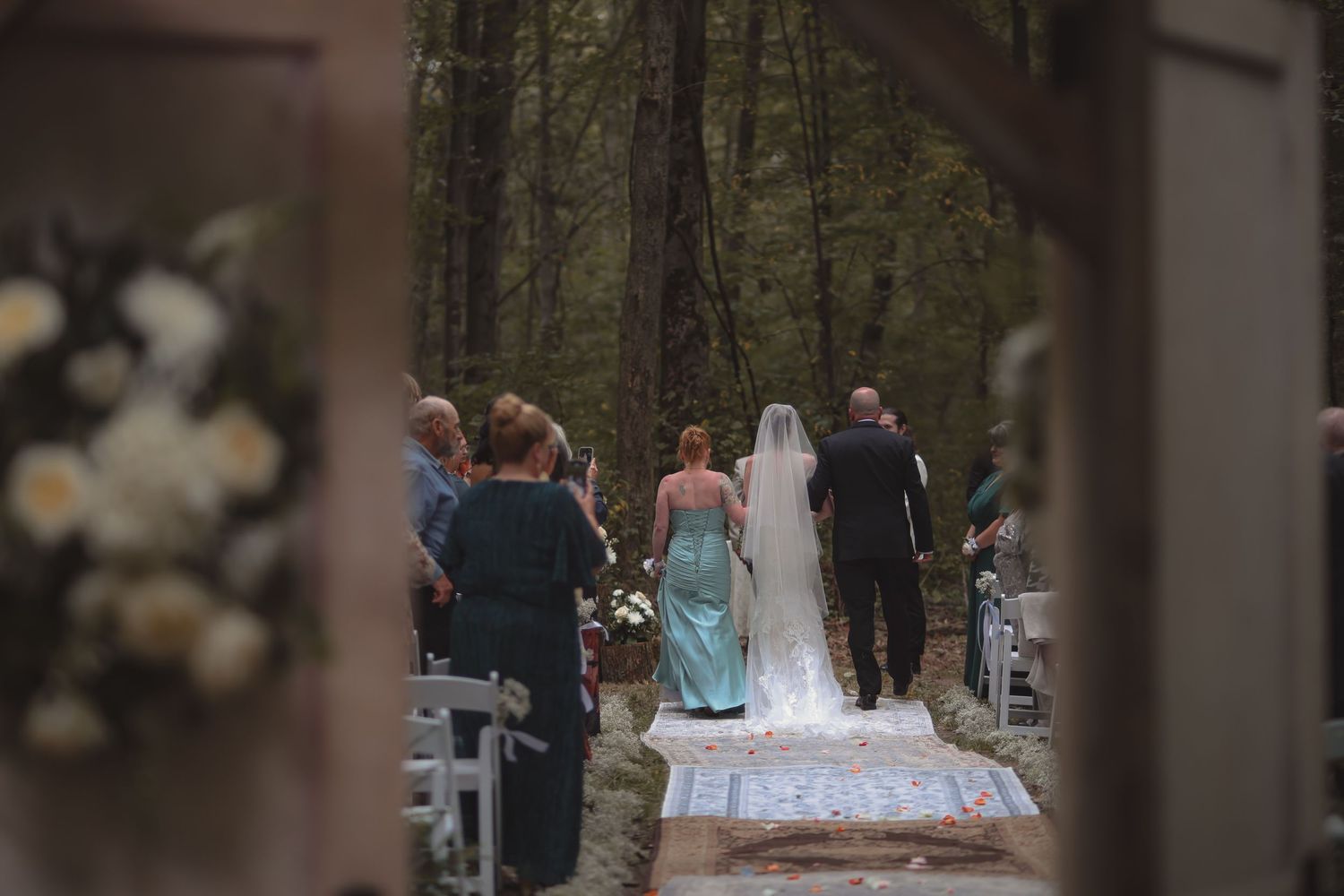 Bride in white gown with veil walks down rustic outdoor forest wedding aisle with escort.