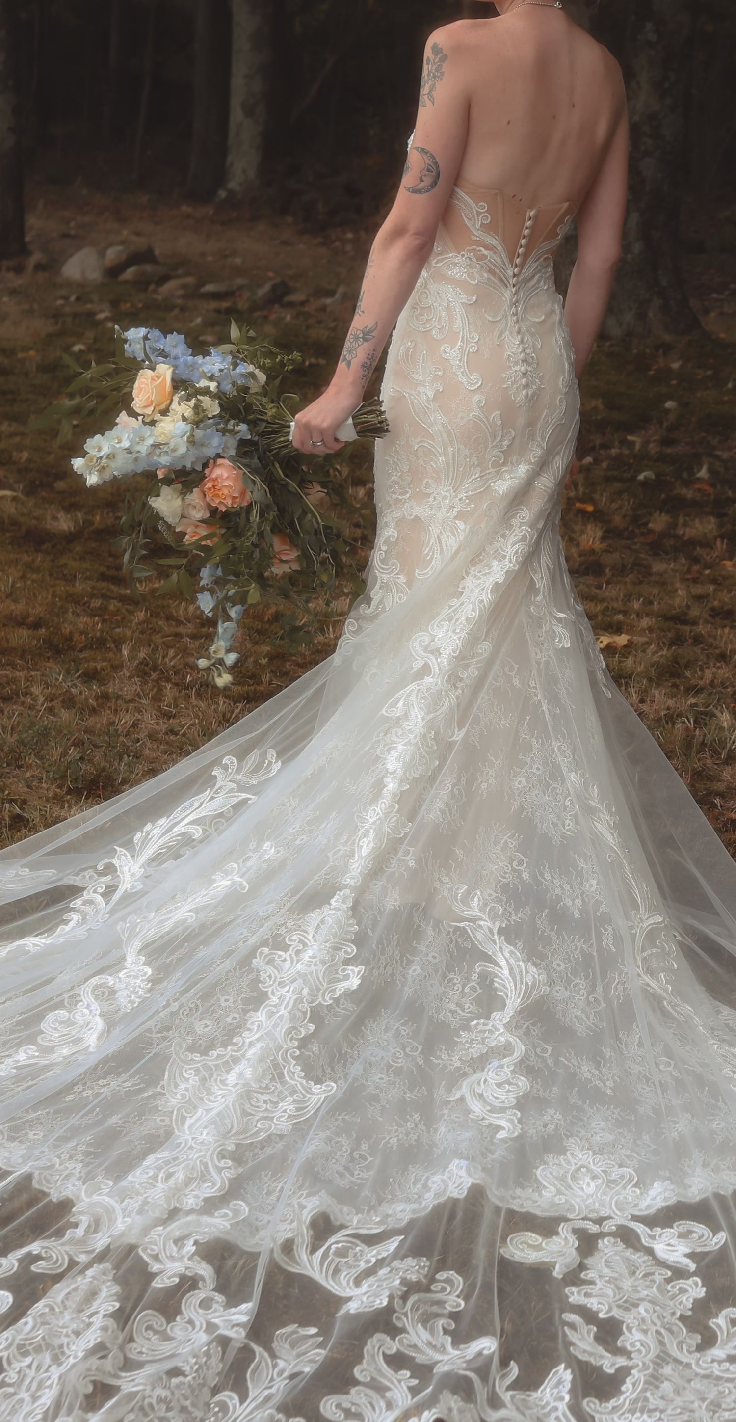 Bride in elegant lace mermaid wedding gown with open back and train, holding pastel bouquet outdoors.