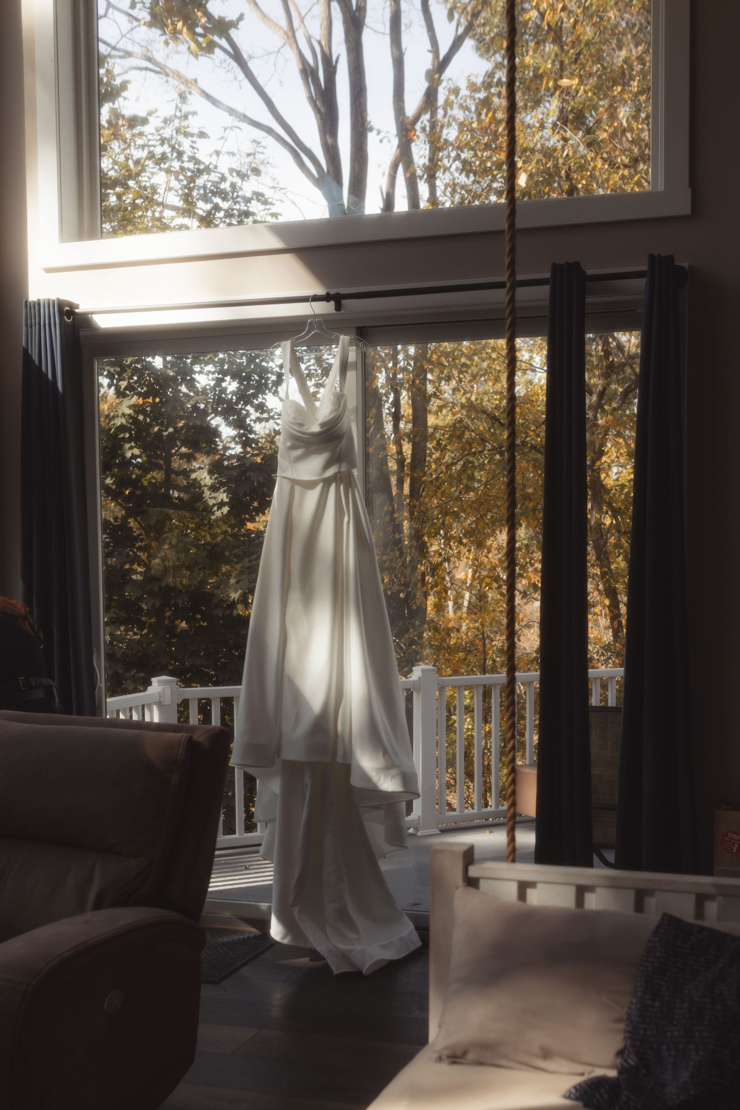 White wedding dress hanging in sunlit window with autumn trees visible outside.