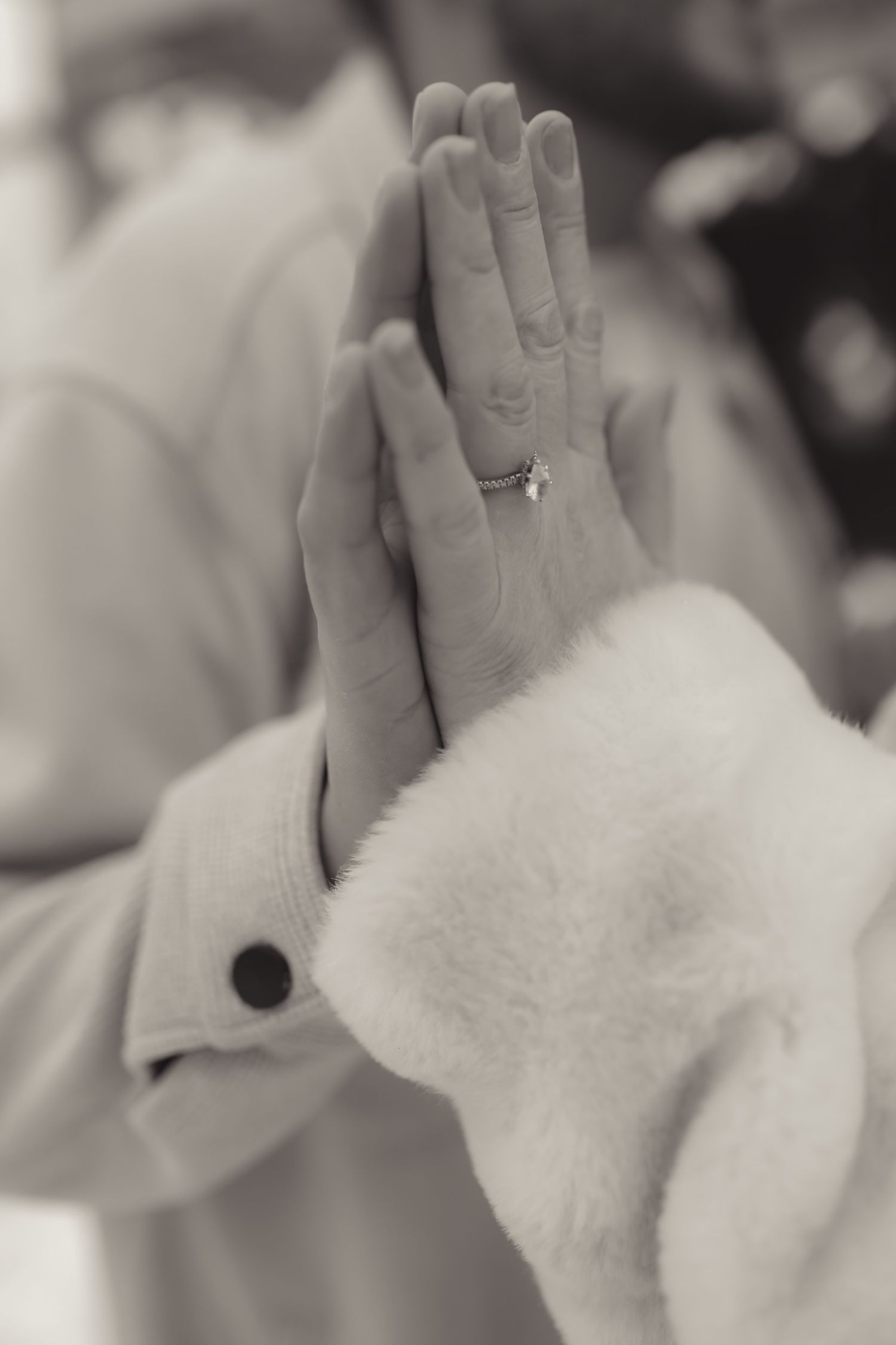 Black and white photo of hands pressed together in prayer, wearing a fur-cuffed coat sleeve and a delicate ring.