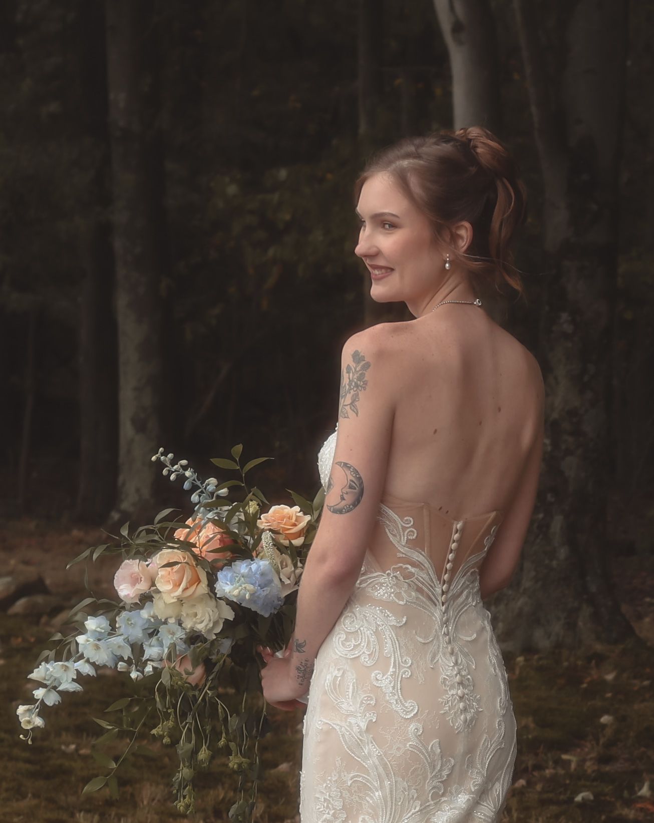 Bride in backless lace wedding gown holds pastel bouquet of roses and blue flowers, smiling in a forest setting.