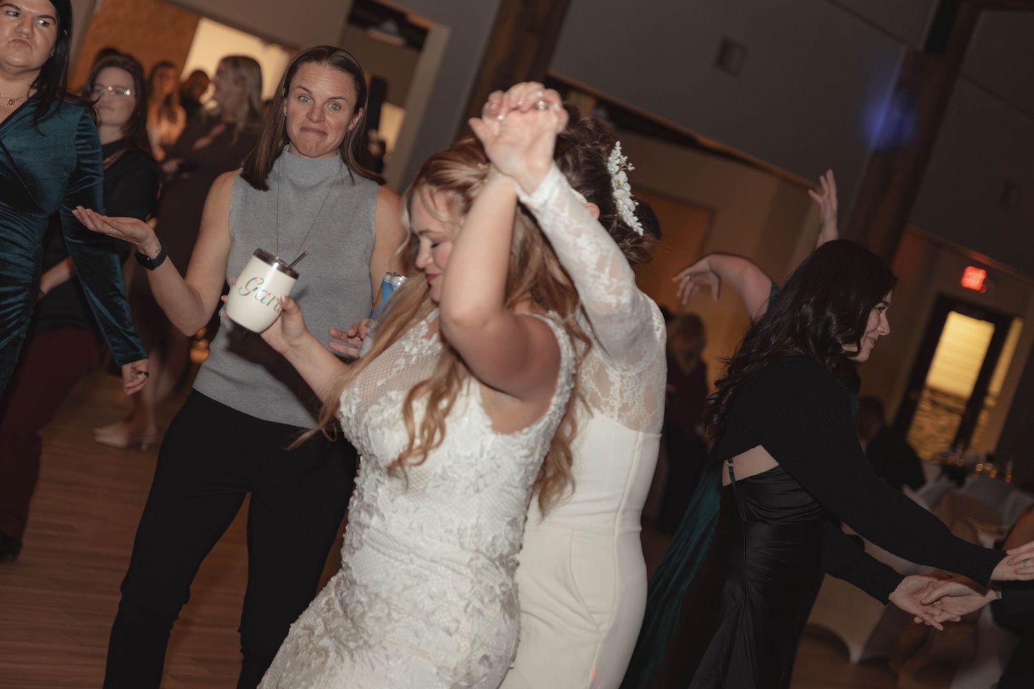 A bride in a white lace gown dances joyfully with guests at a lively wedding reception.