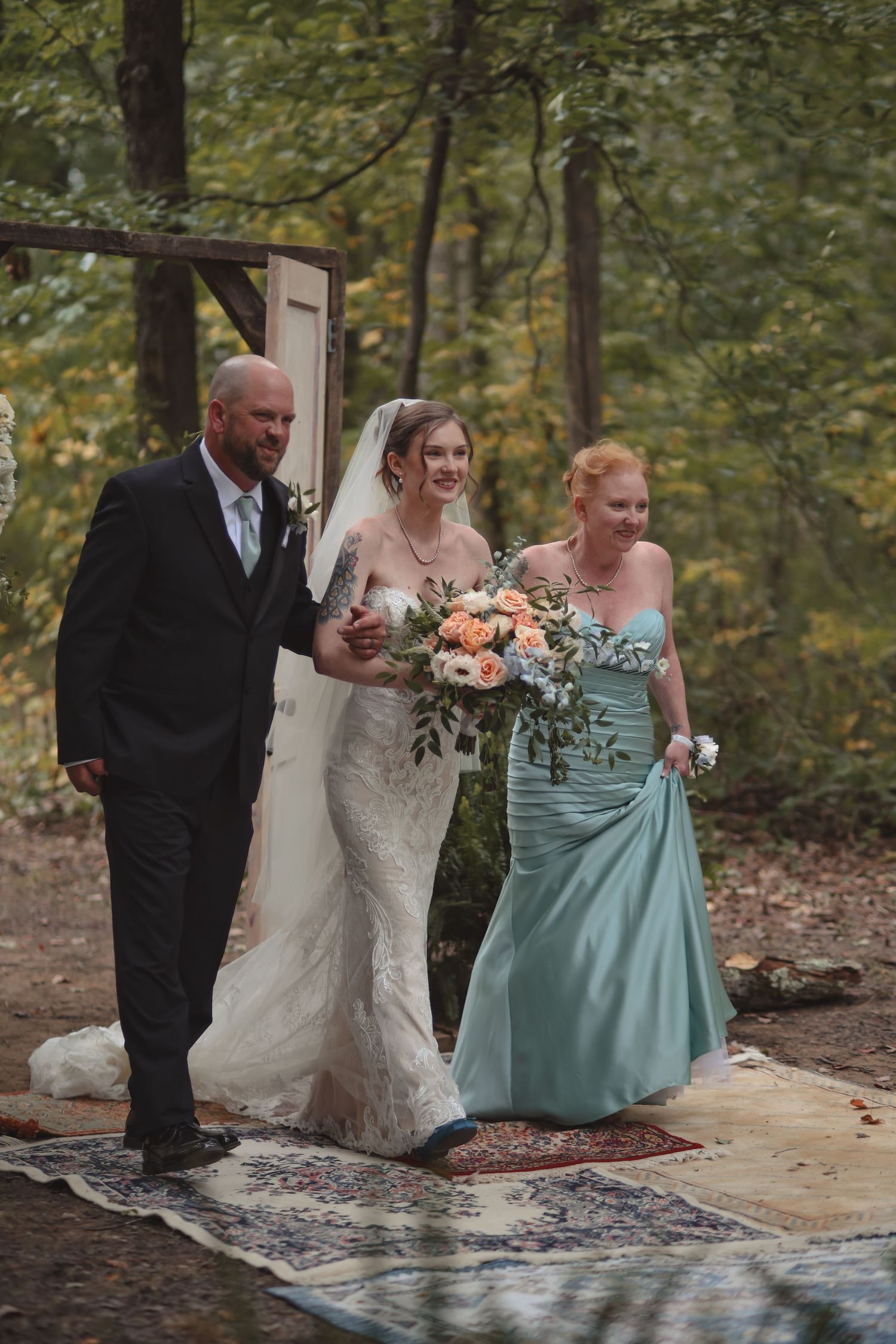 Bride walks down forest aisle in lace gown with veil, escorted by father and bridesmaid in teal.