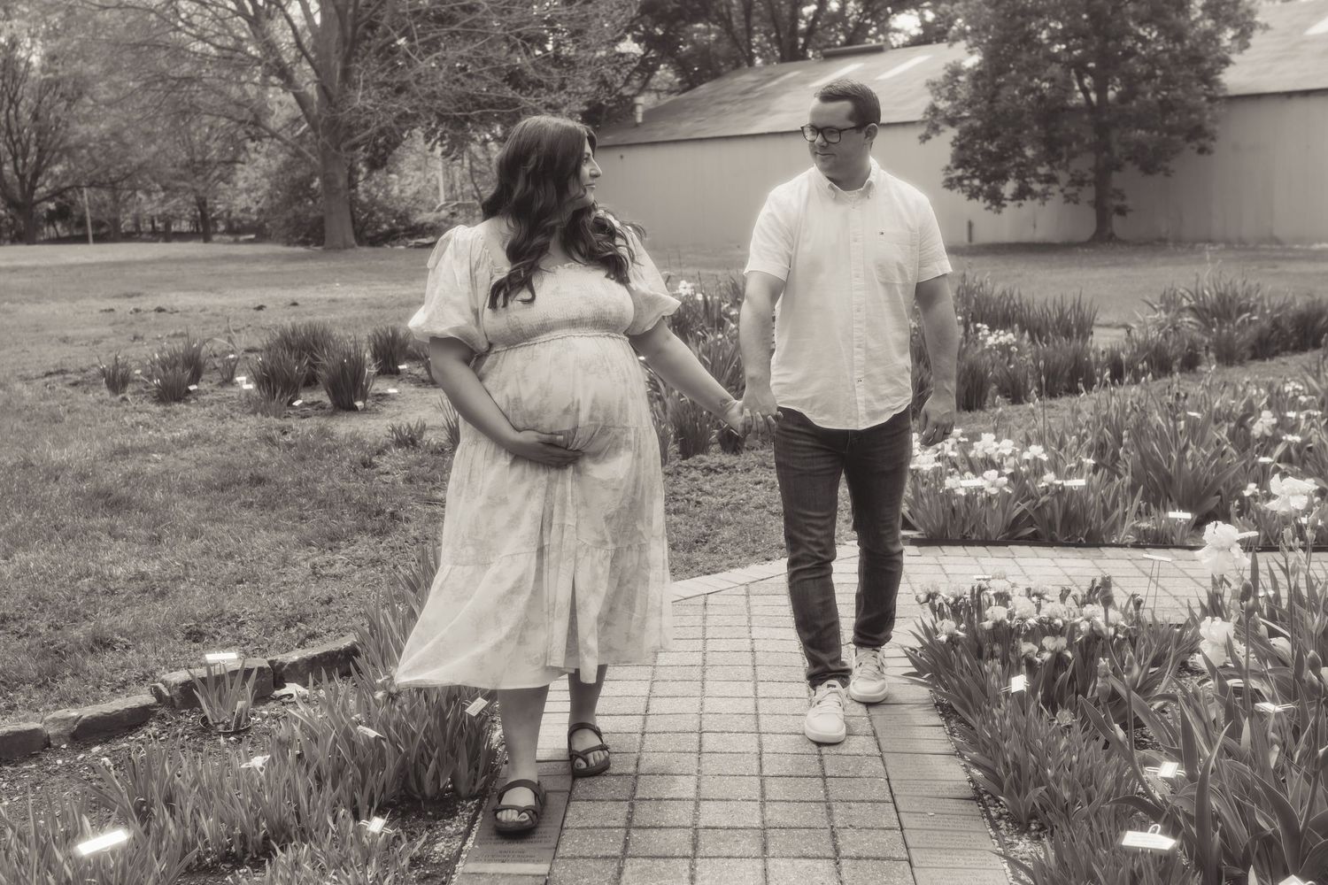 Black and white photo of a pregnant woman in a white dress holding hands with a man on a garden pathway.