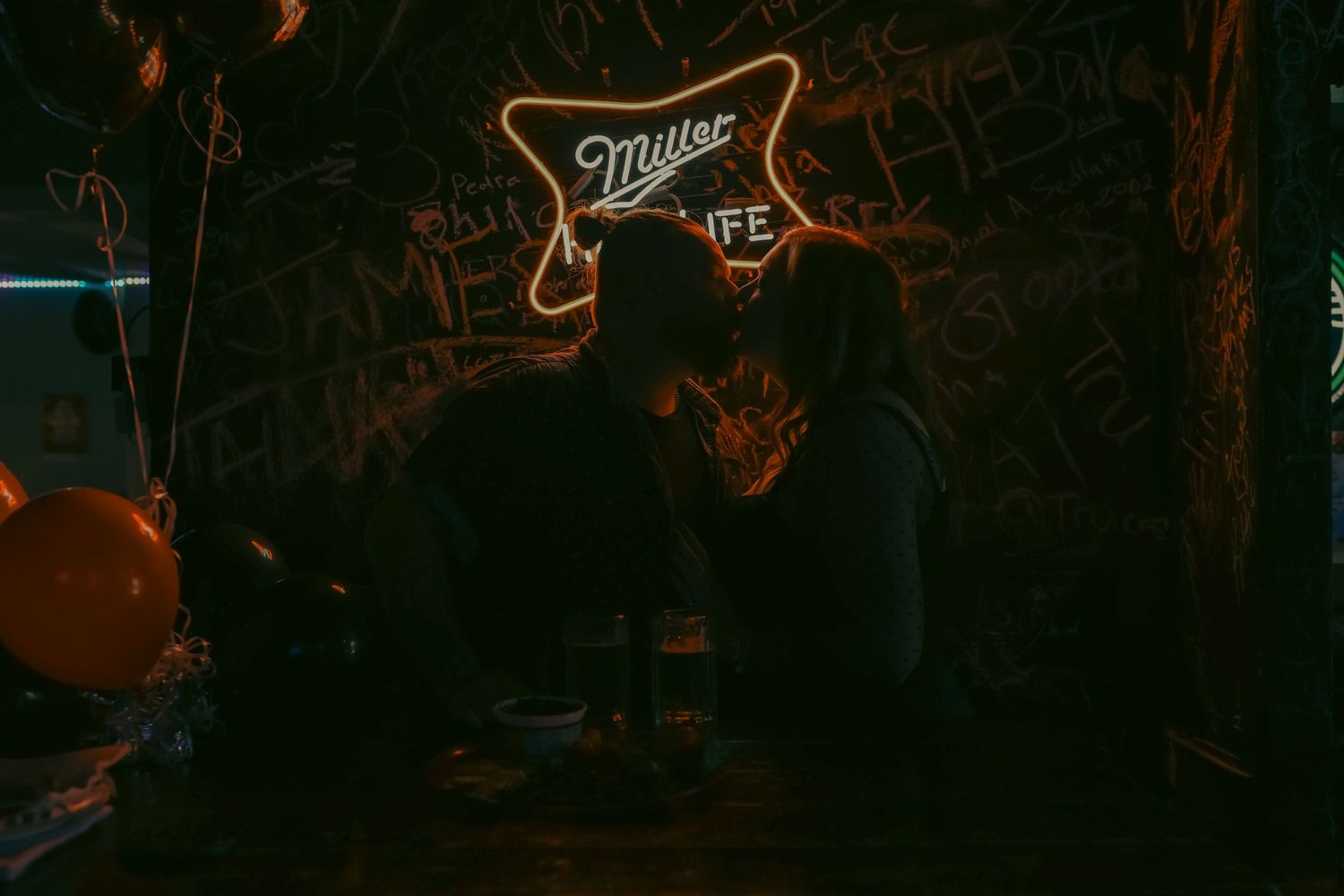 Couple sharing a romantic kiss under a glowing Miller Lite neon sign in a dark bar.