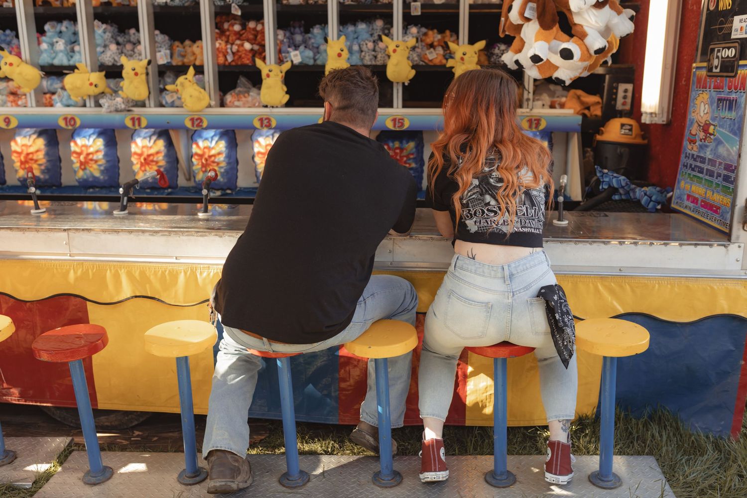 Couple sitting on stools at a carnival game booth filled with Pikachu plush prizes.