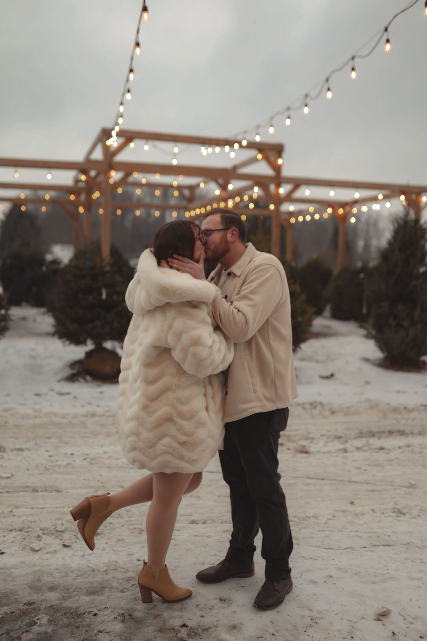 Couple kissing at a snowy Christmas tree farm under a wooden pergola with string lights.