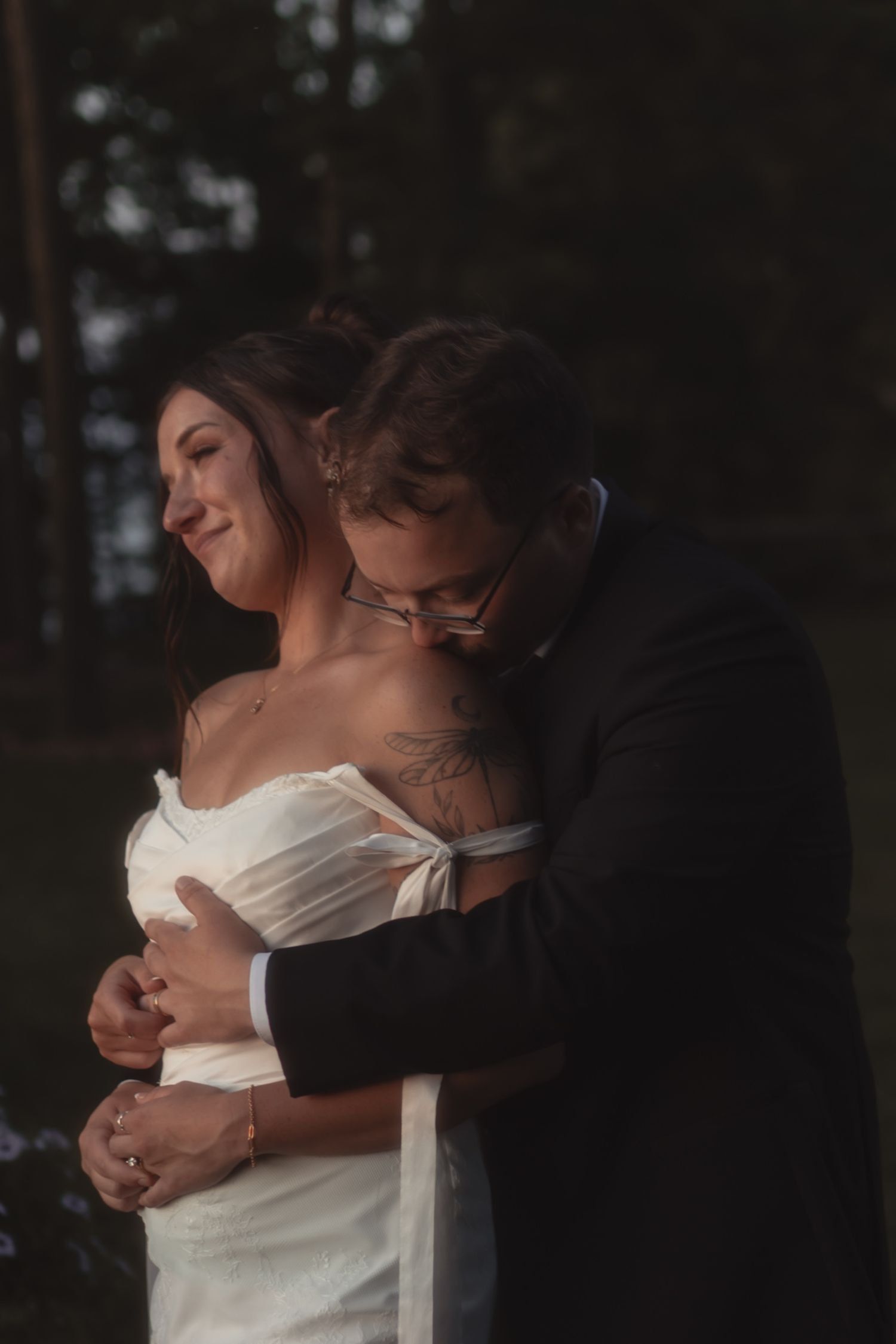 Groom in black suit embraces bride in white off-shoulder dress, sharing a tender moment outdoors at their wedding.