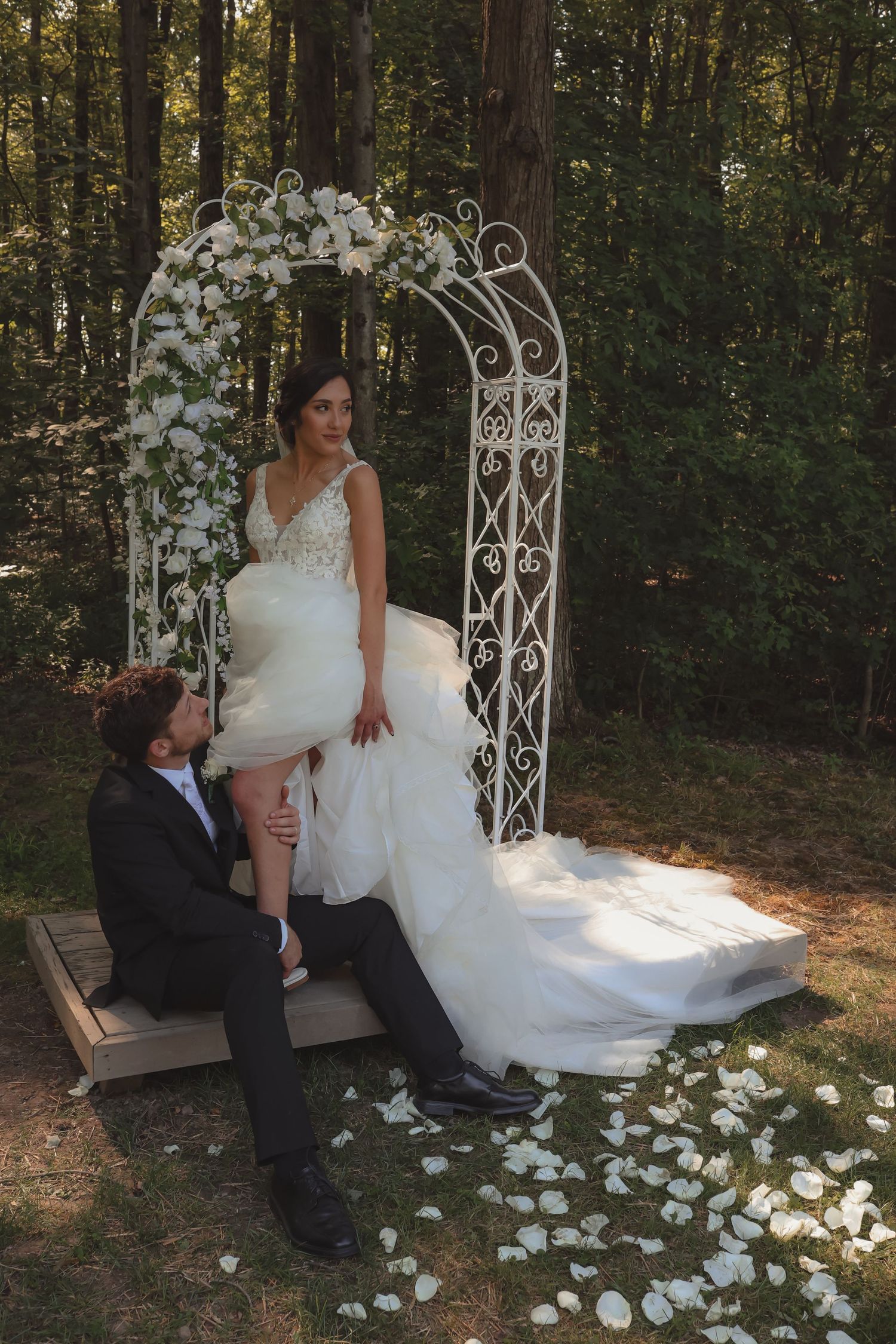 Bride and groom pose at a floral white iron arch in a forest wedding venue with rose petals scattered below.