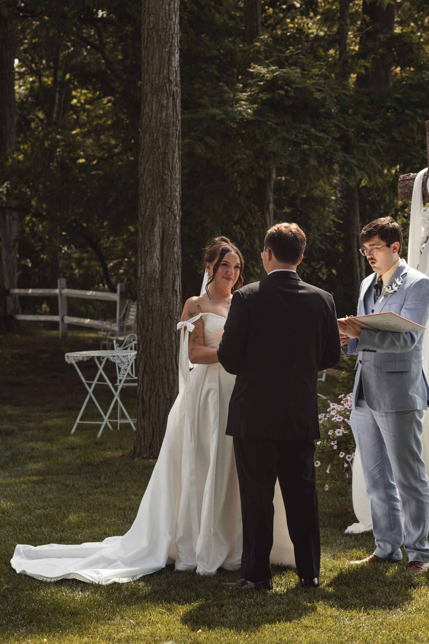 Bride in white gown and groom in tuxedo exchange vows outdoors with officiant in gray suit amid lush green trees.