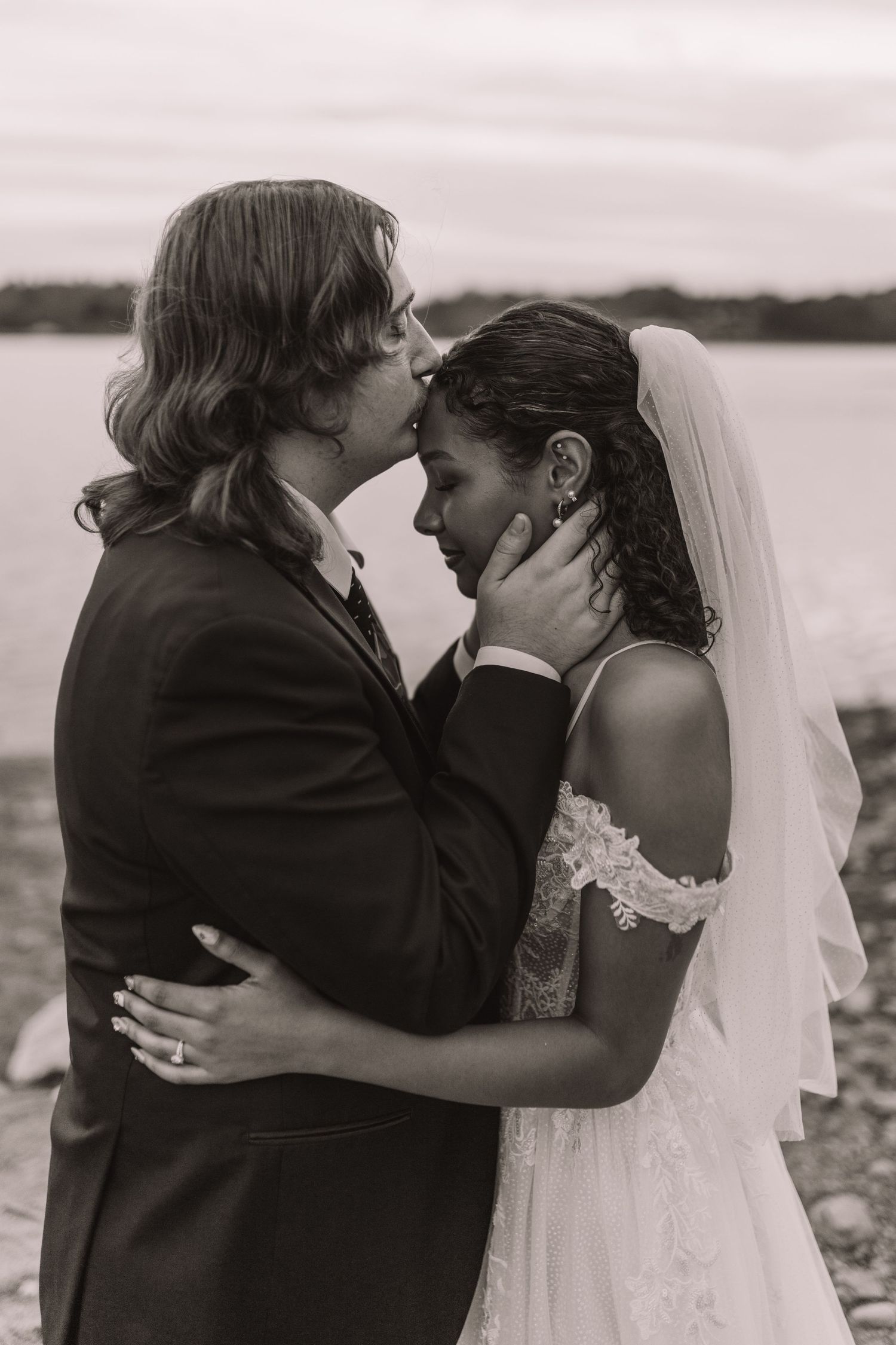 Black and white photo of a groom tenderly holding bride's face for a kiss near a waterfront.