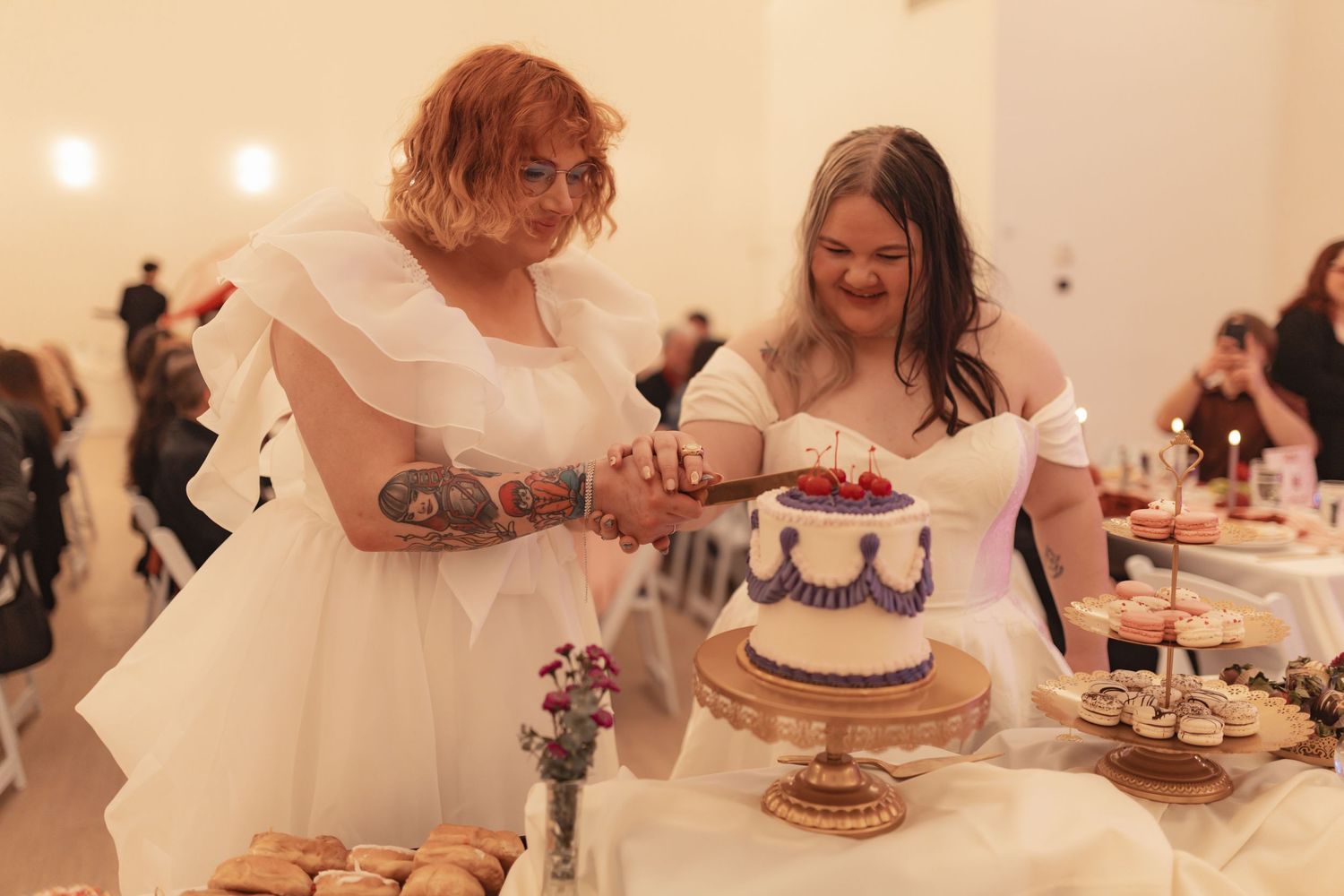 Two brides in white wedding dresses smile while cutting a decorated white and purple wedding cake.