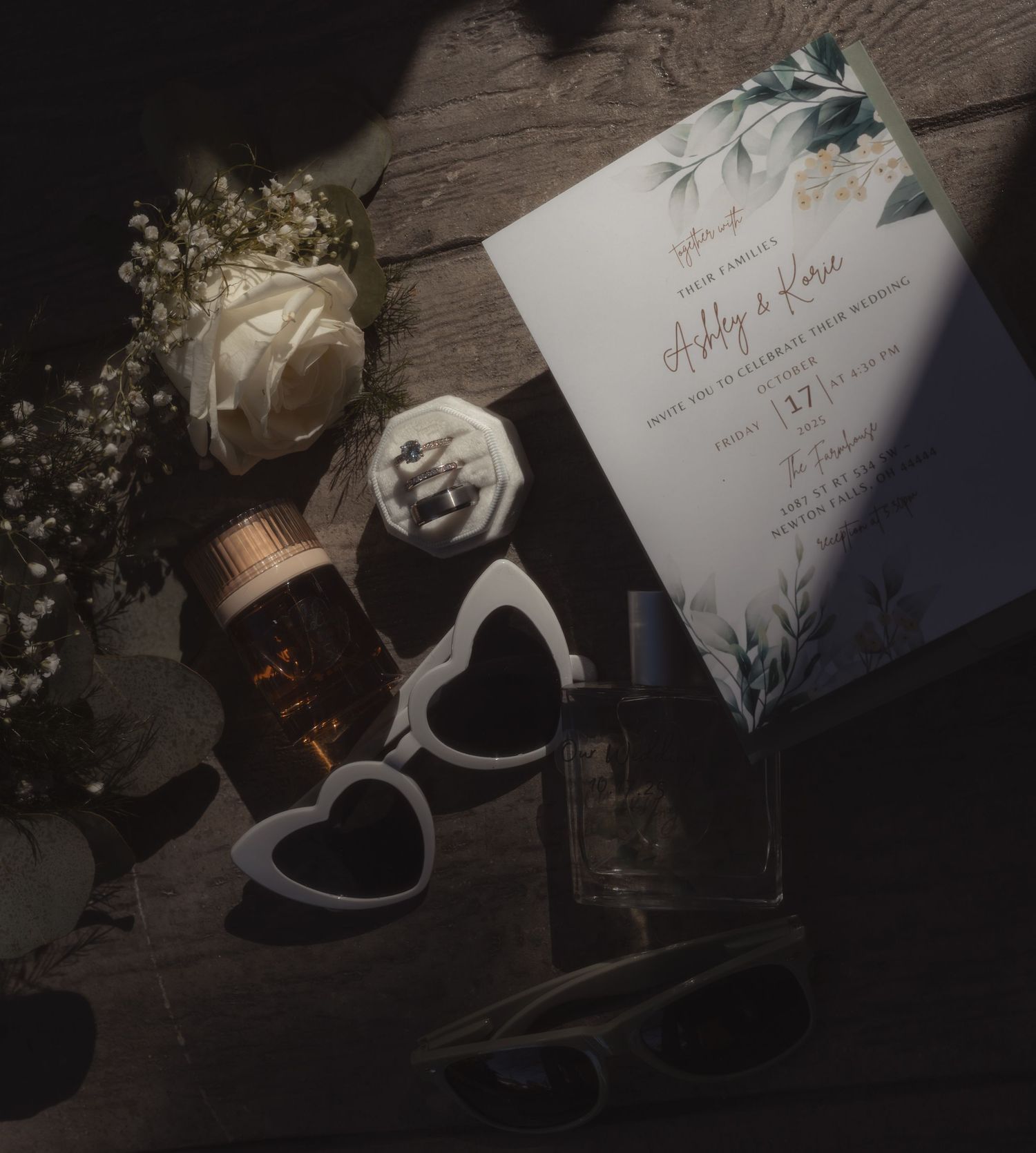 Wedding flat lay with floral invitation, white rose, heart sunglasses, perfume, and ring box on rustic wood.