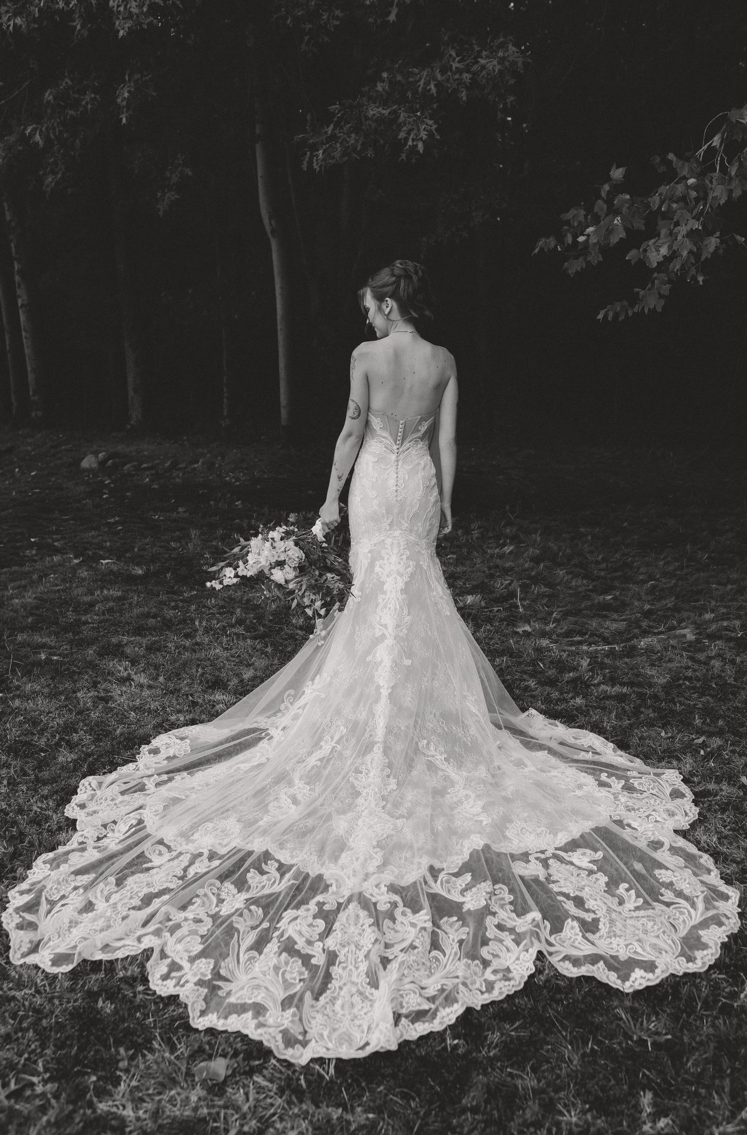 Bride in elegant backless lace mermaid wedding gown with long train, holding bouquet in black and white photo.