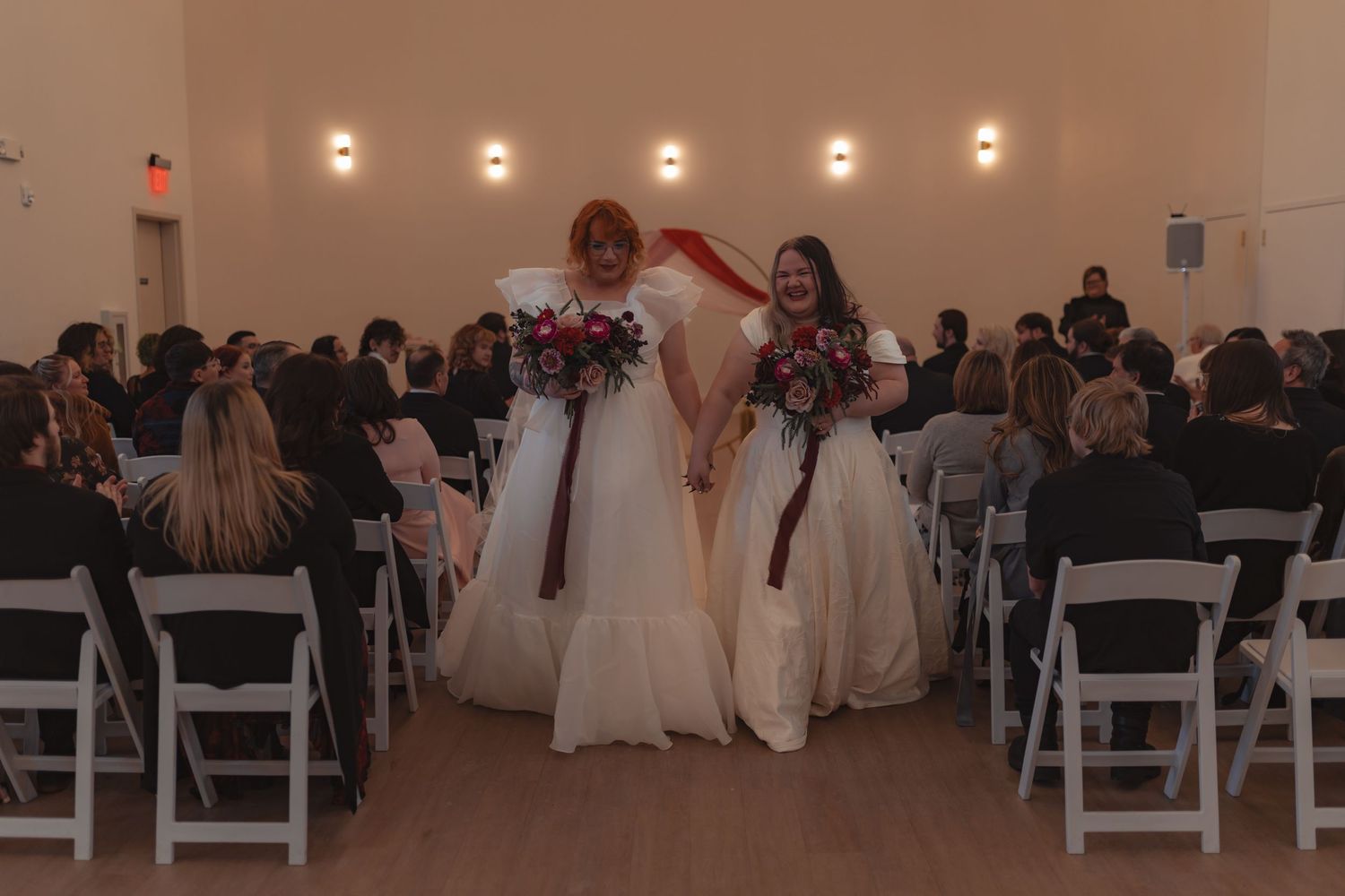 Two brides in white gowns walk down the aisle holding bouquets at their wedding ceremony.