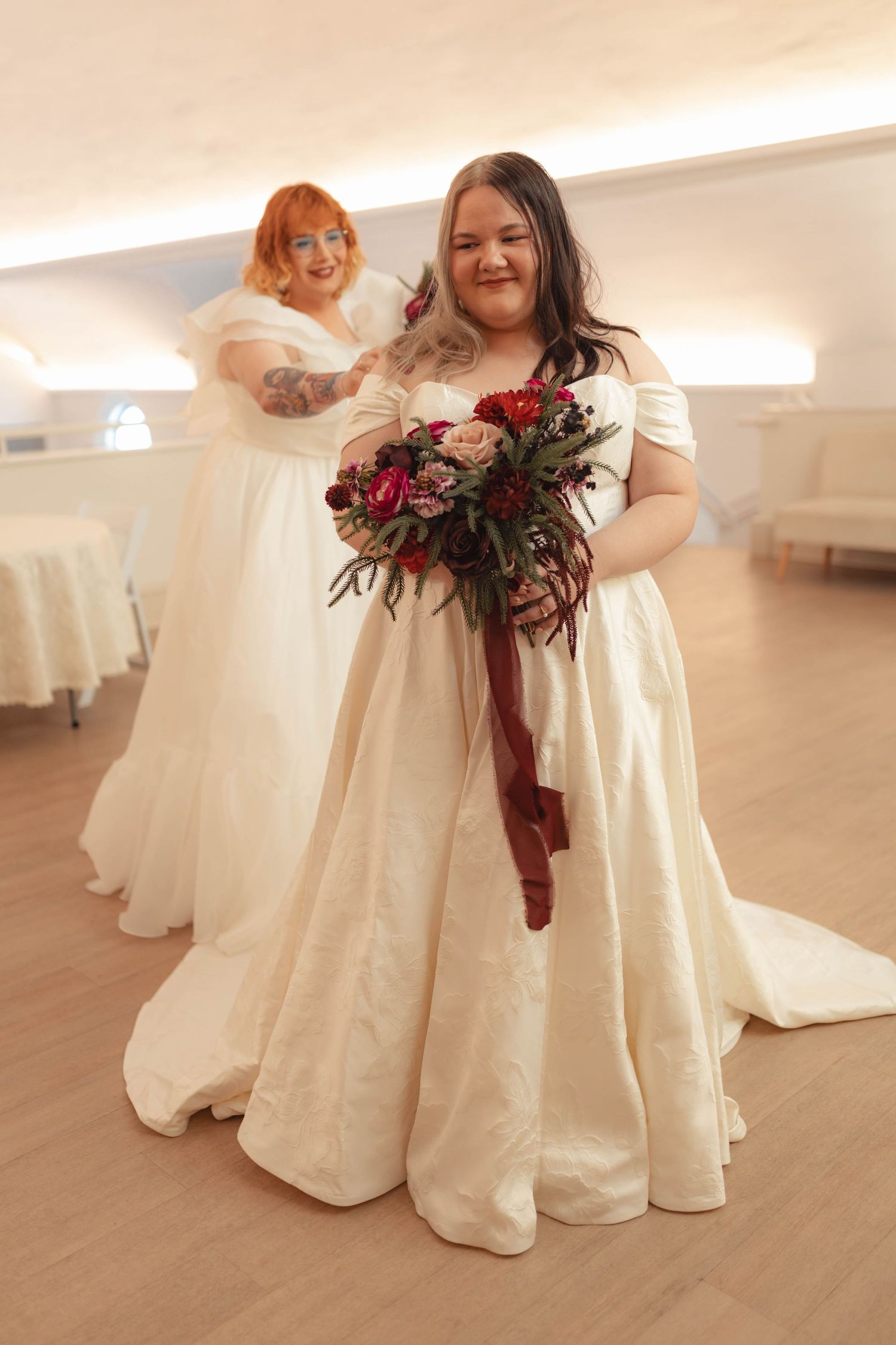 Two plus-size brides in white gowns, one holding a rich red and pink floral bouquet indoors.