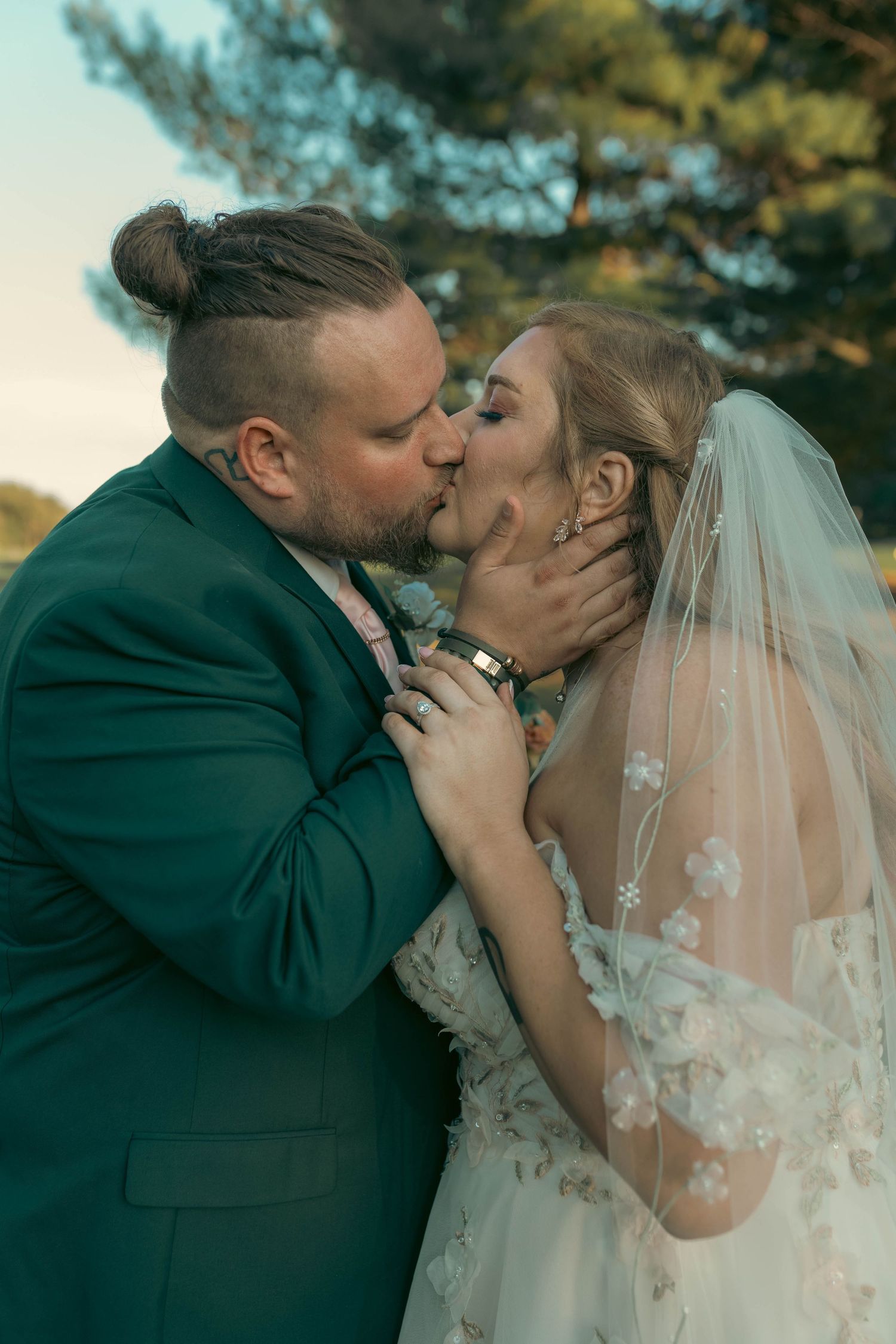 Bride in floral veil and groom in green suit share a tender kiss outdoors on their wedding day.