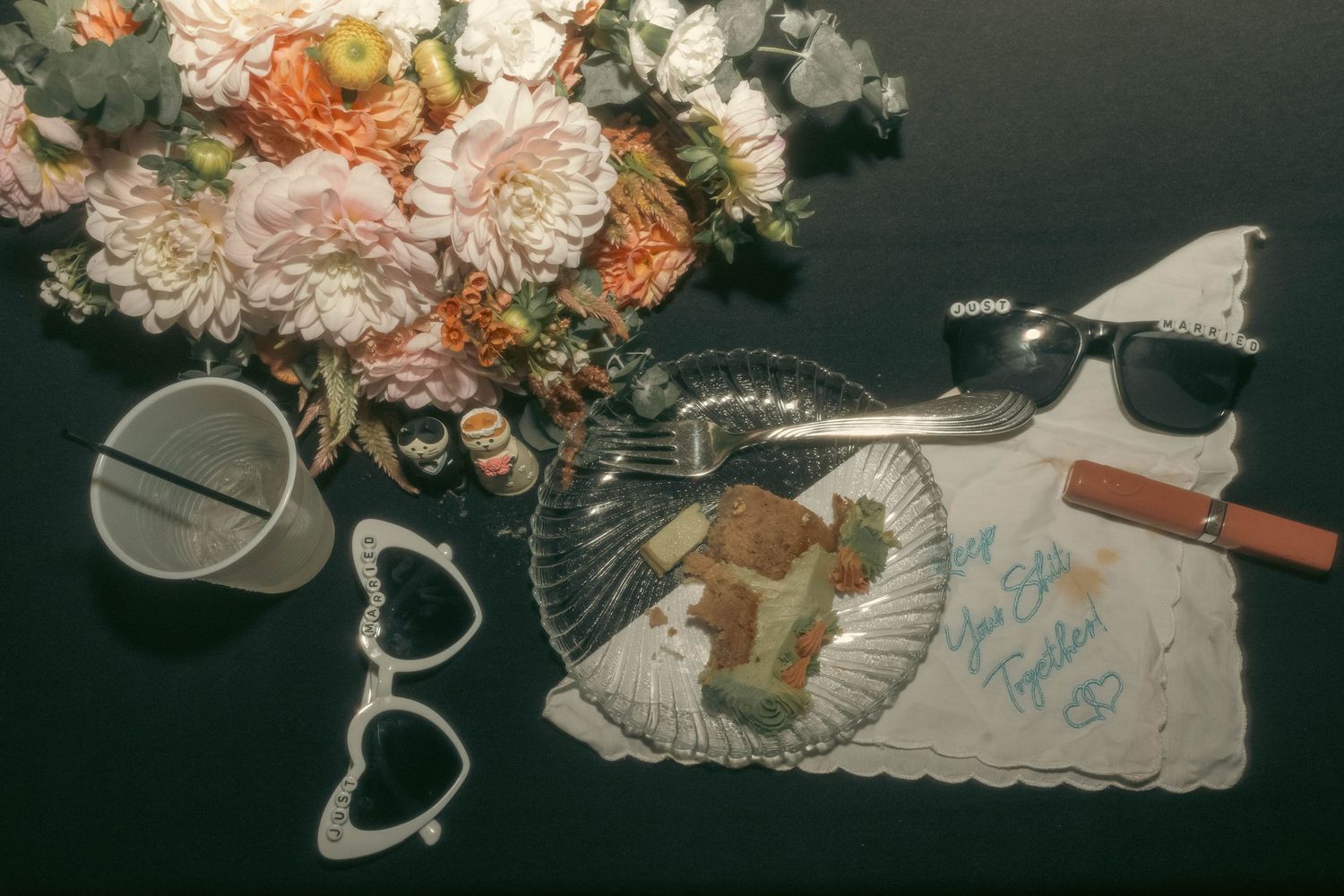 Flat lay of pink flowers, sunglasses, fork, heart glasses, and a signed napkin on a dark surface.
