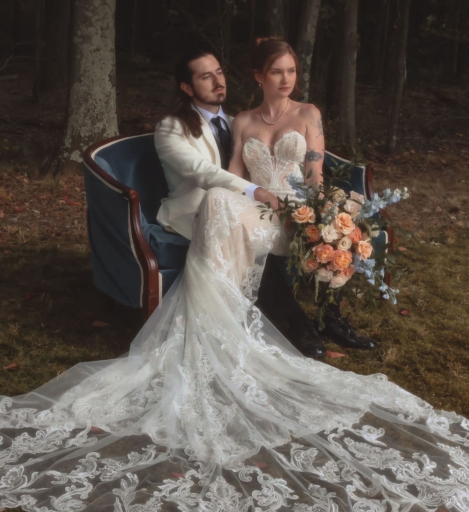 Bride in lace mermaid gown and groom in white suit pose with floral bouquet in a woodland wedding setting.