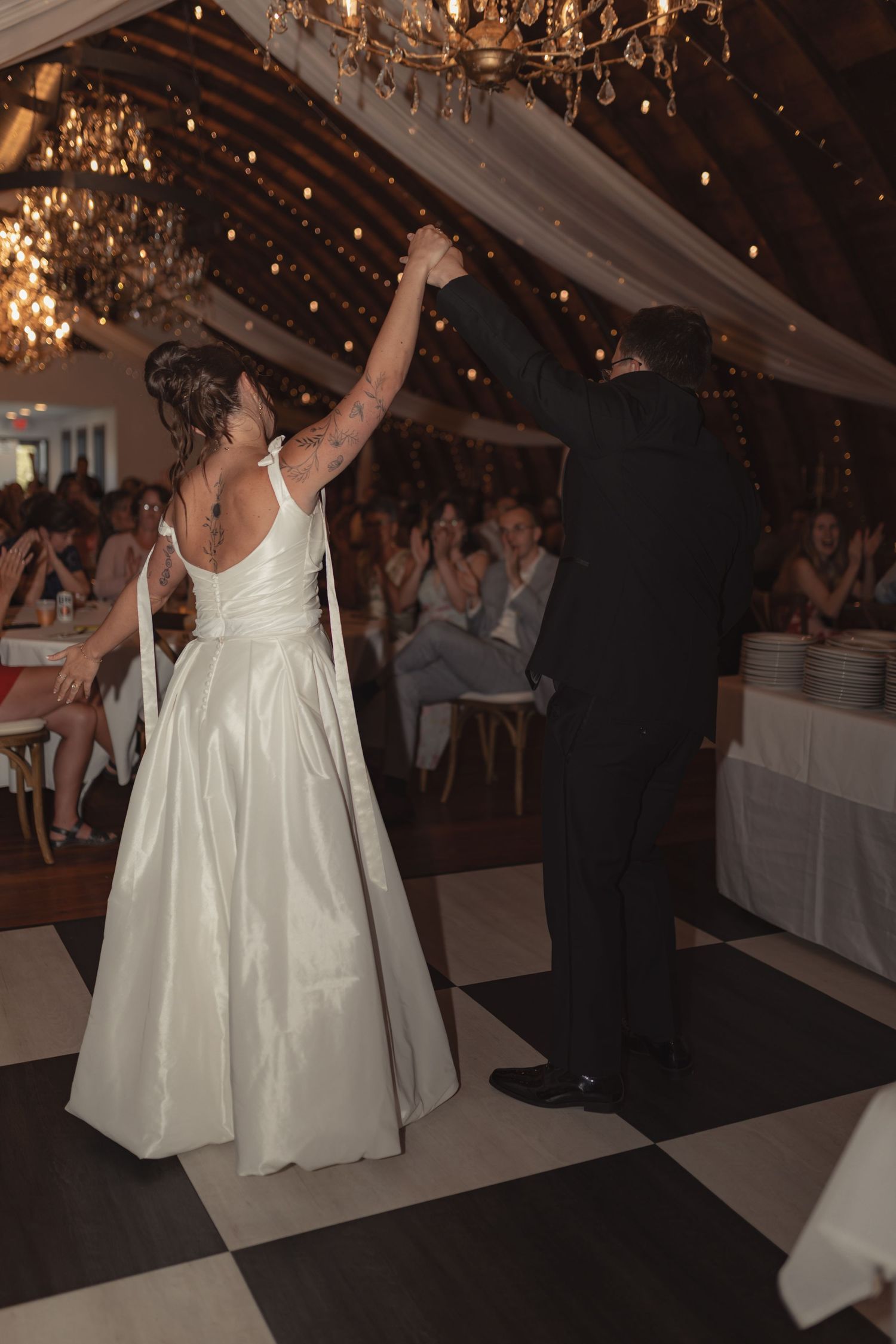 Bride and groom share first dance on checkered floor in elegant chandelier-lit barn wedding reception.