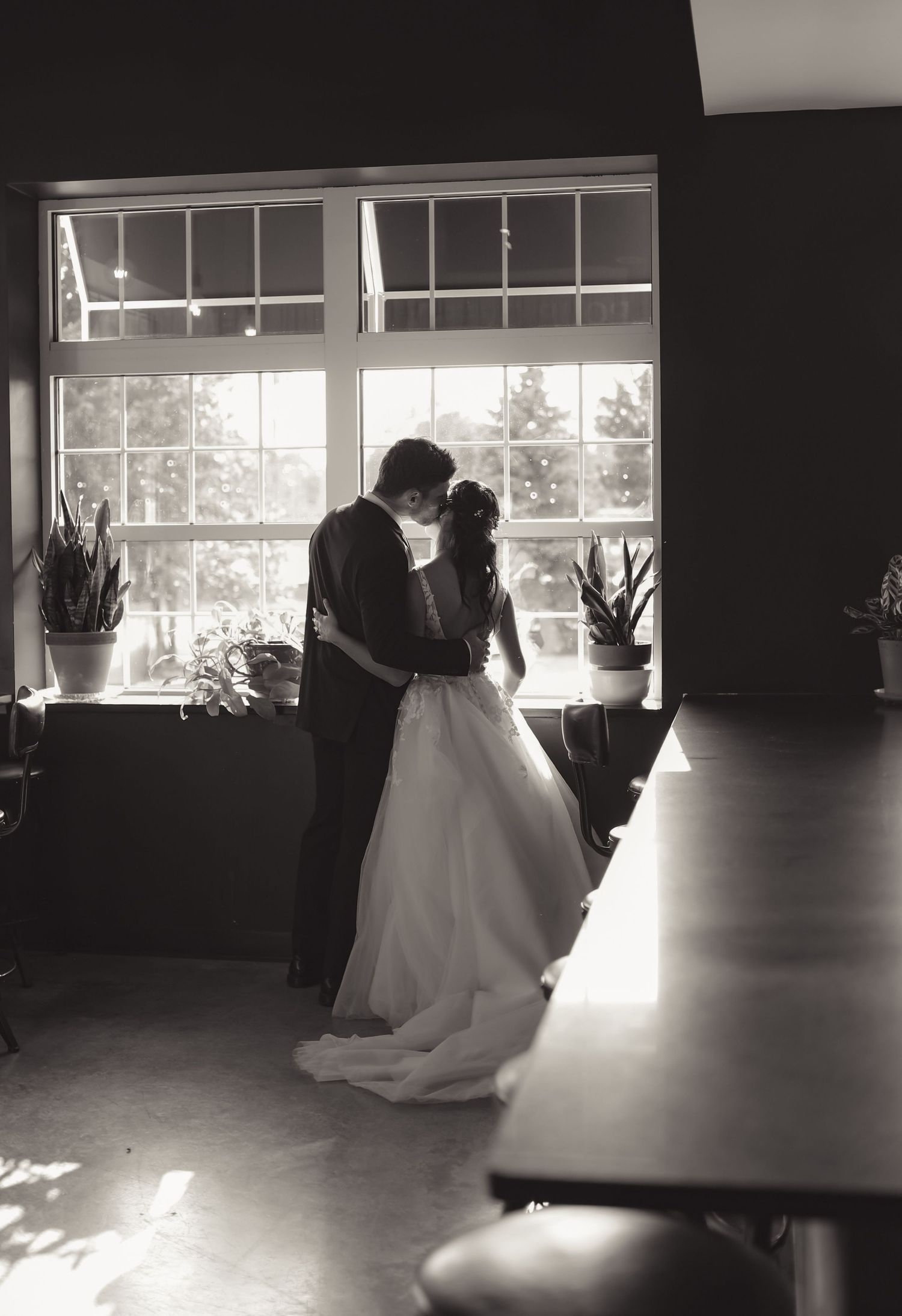Bride and groom share intimate embrace by large window in black and white photo.