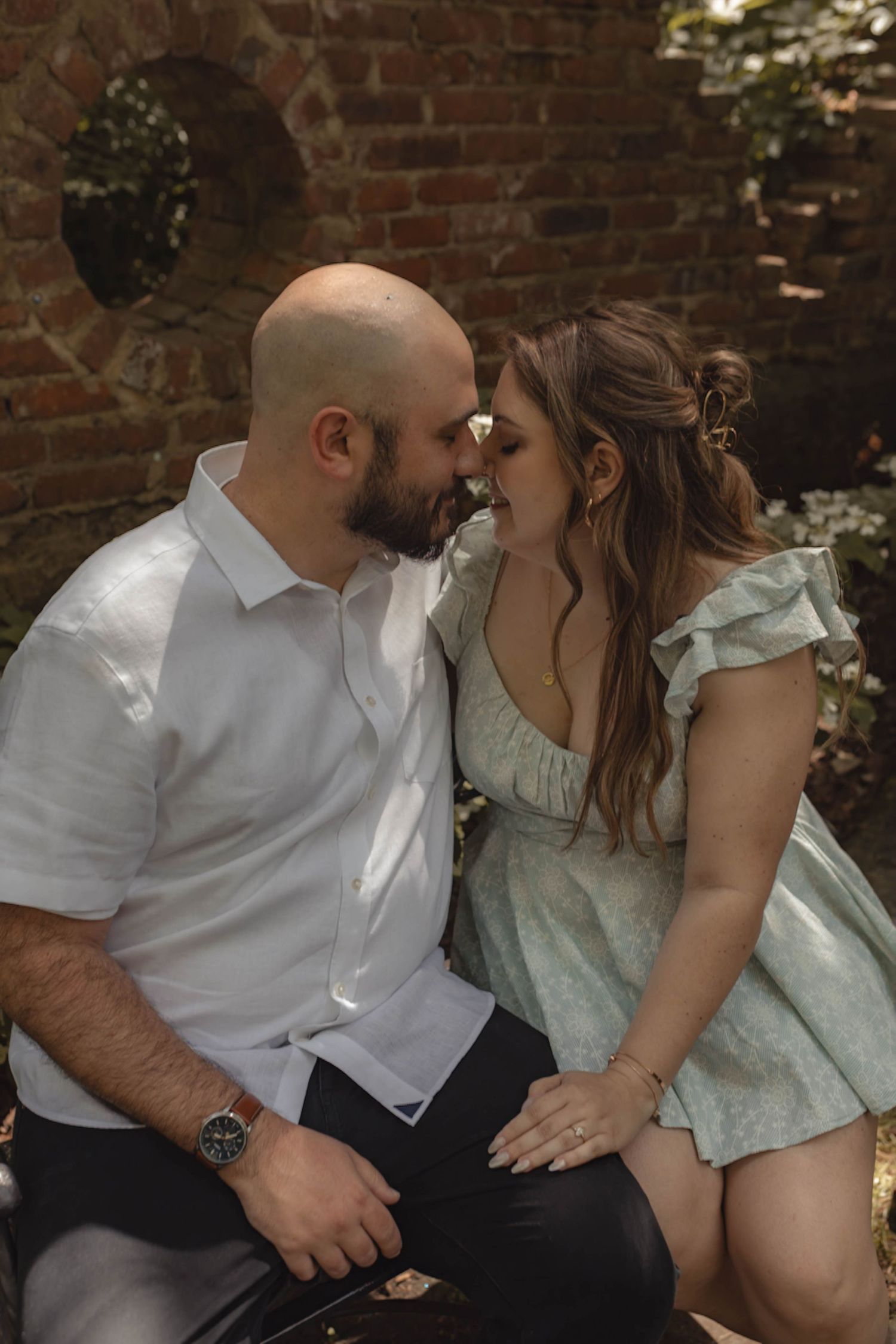 A couple shares a romantic moment, leaning in close while seated against a rustic brick wall outdoors.