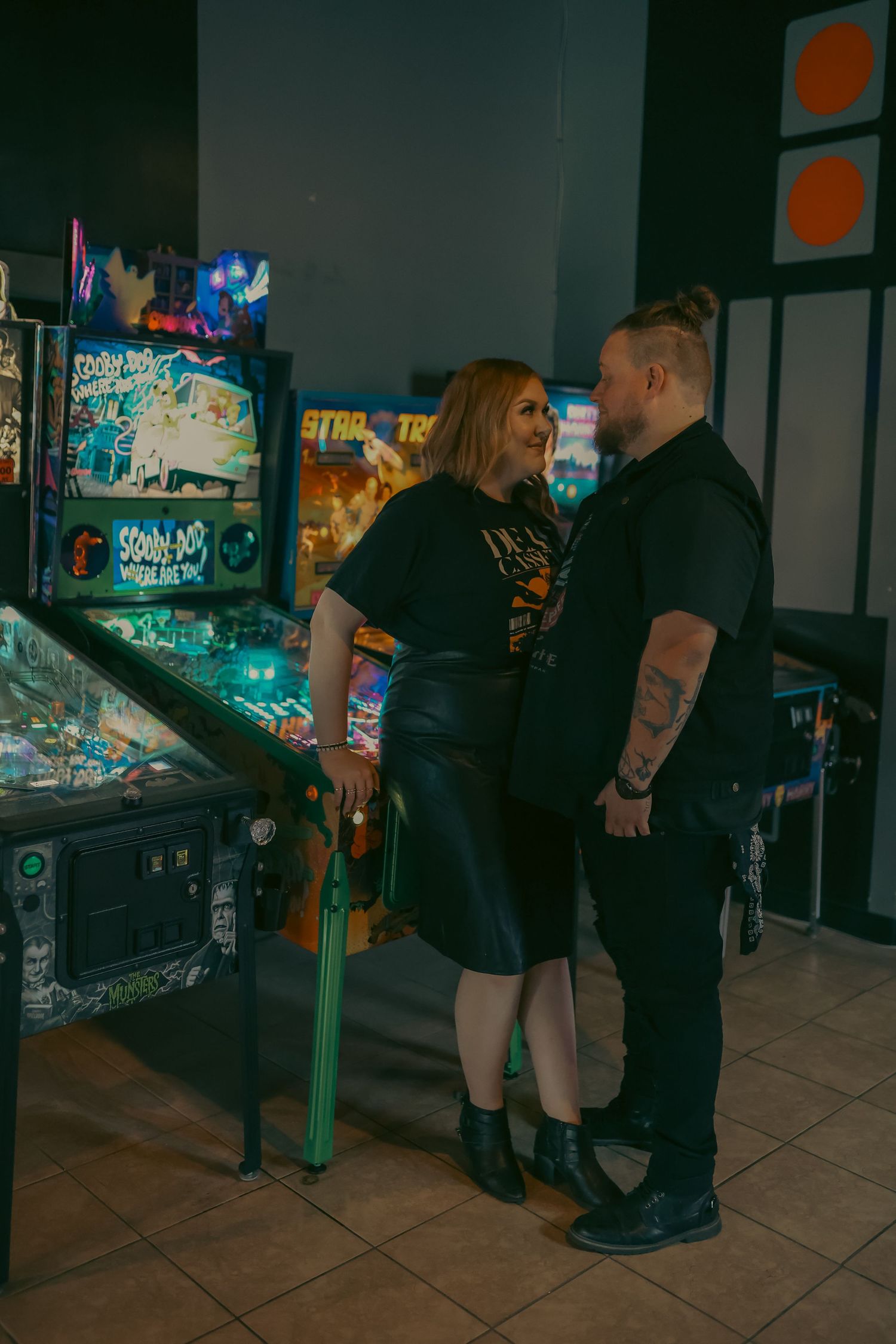 Couple smiling at each other beside vintage pinball machines in an arcade game room.