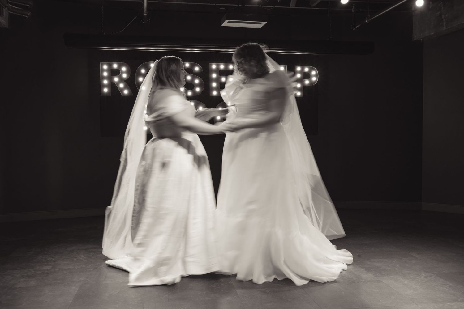 Two brides in white wedding gowns and veils share an intimate moment on a dark stage with marquee lights.