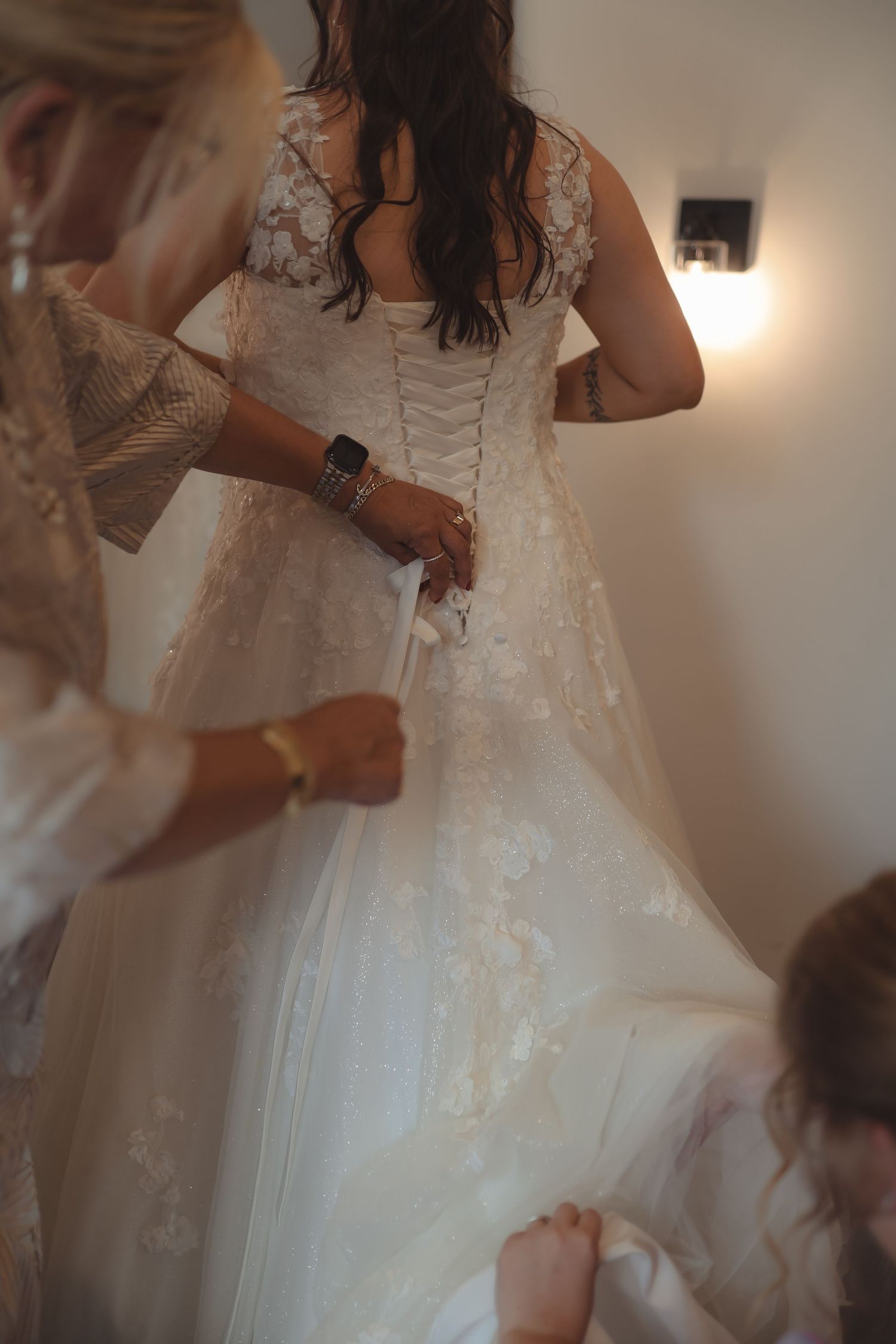 Bridesmaids helping bride lace up elegant white lace corset wedding dress before ceremony.