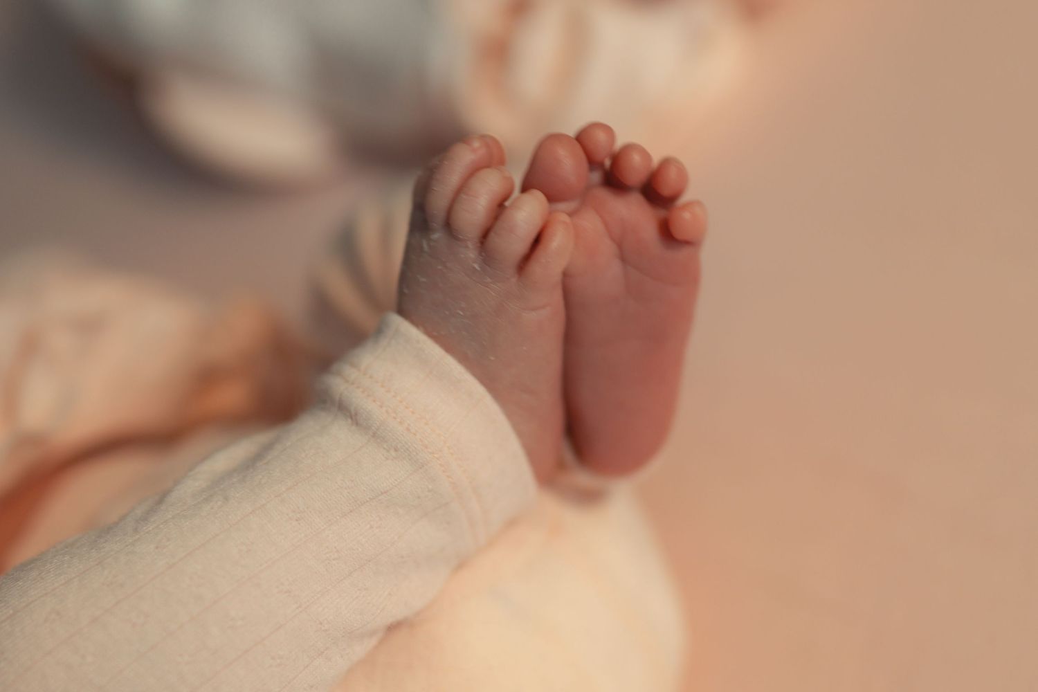 Close-up of a newborn baby's tiny feet wrapped in a soft white blanket on a pink background.