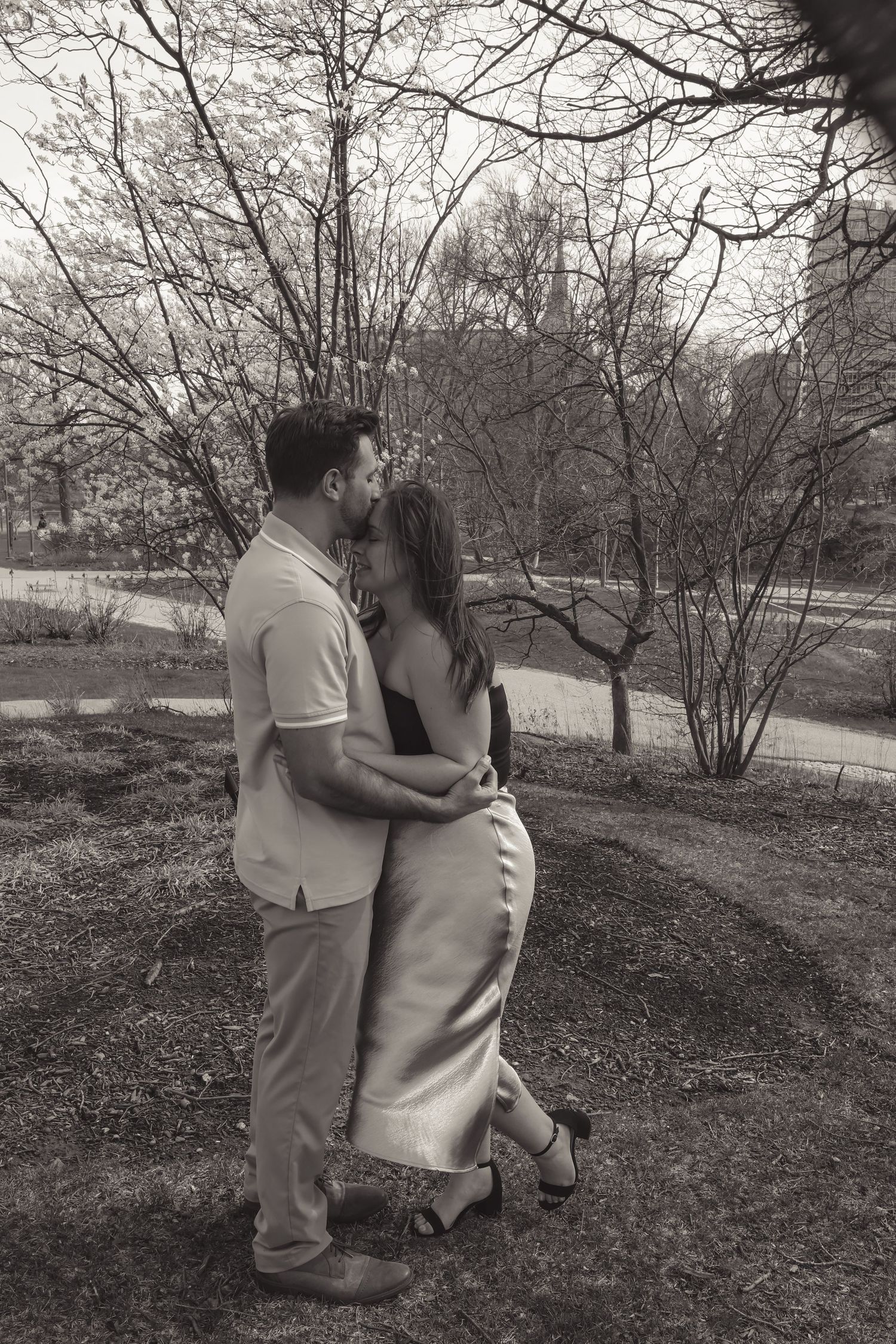 Black and white photo of a couple embracing and kissing in a park with bare trees in the background.