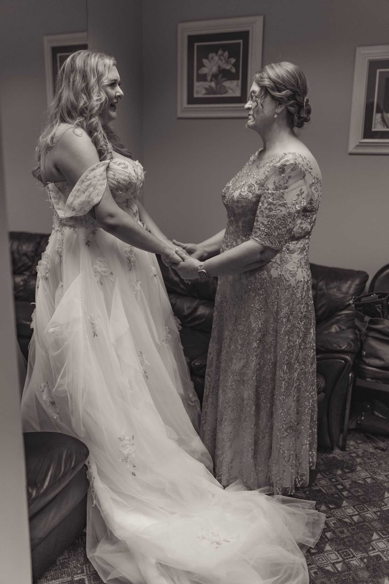 Black and white photo of bride holding hands with woman in lace dress, sharing emotional moment before wedding ceremony.