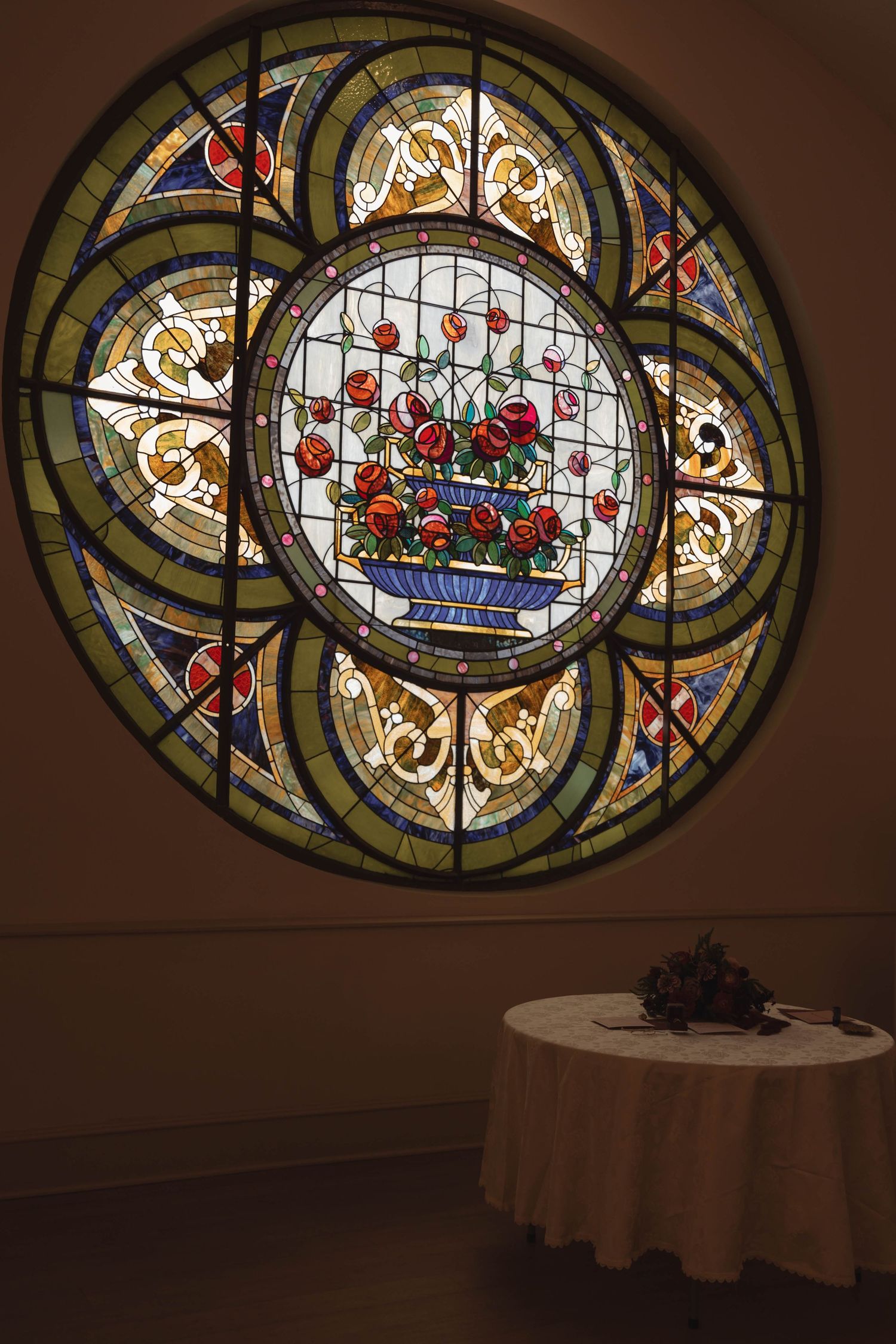 Ornate circular stained glass window featuring a floral bouquet design with red roses, above a small round table in an elegant interior.