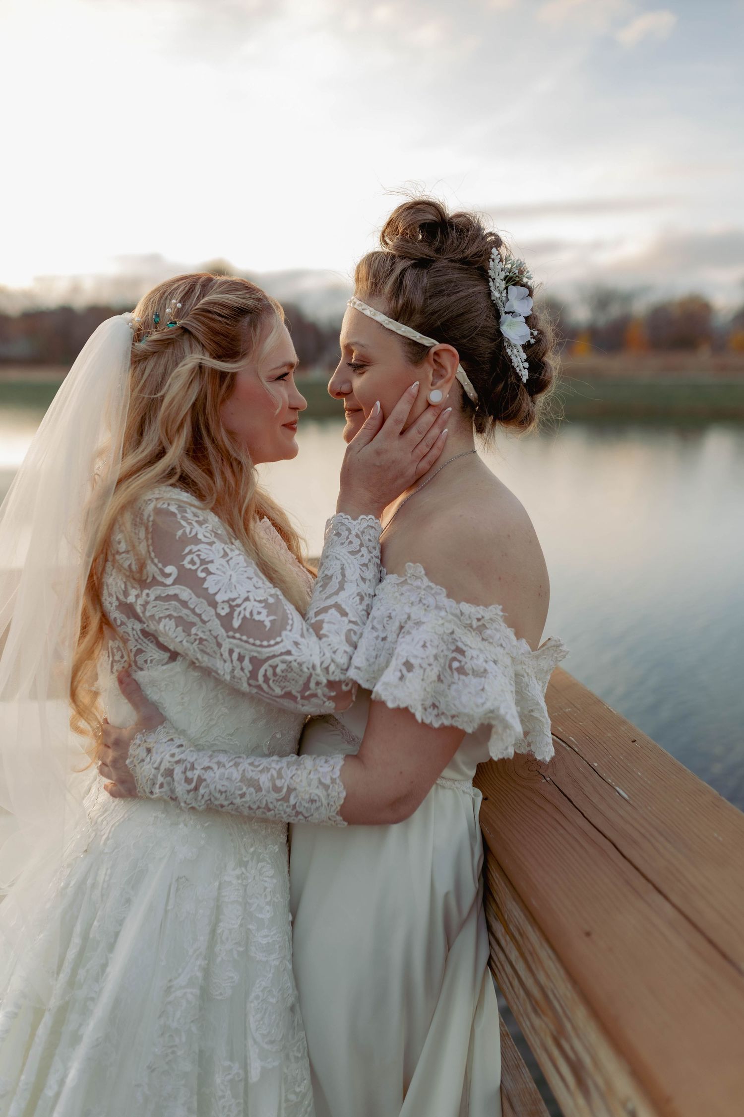Two brides in white wedding gowns share a tender moment on a wooden bridge overlooking a serene lake at sunset.