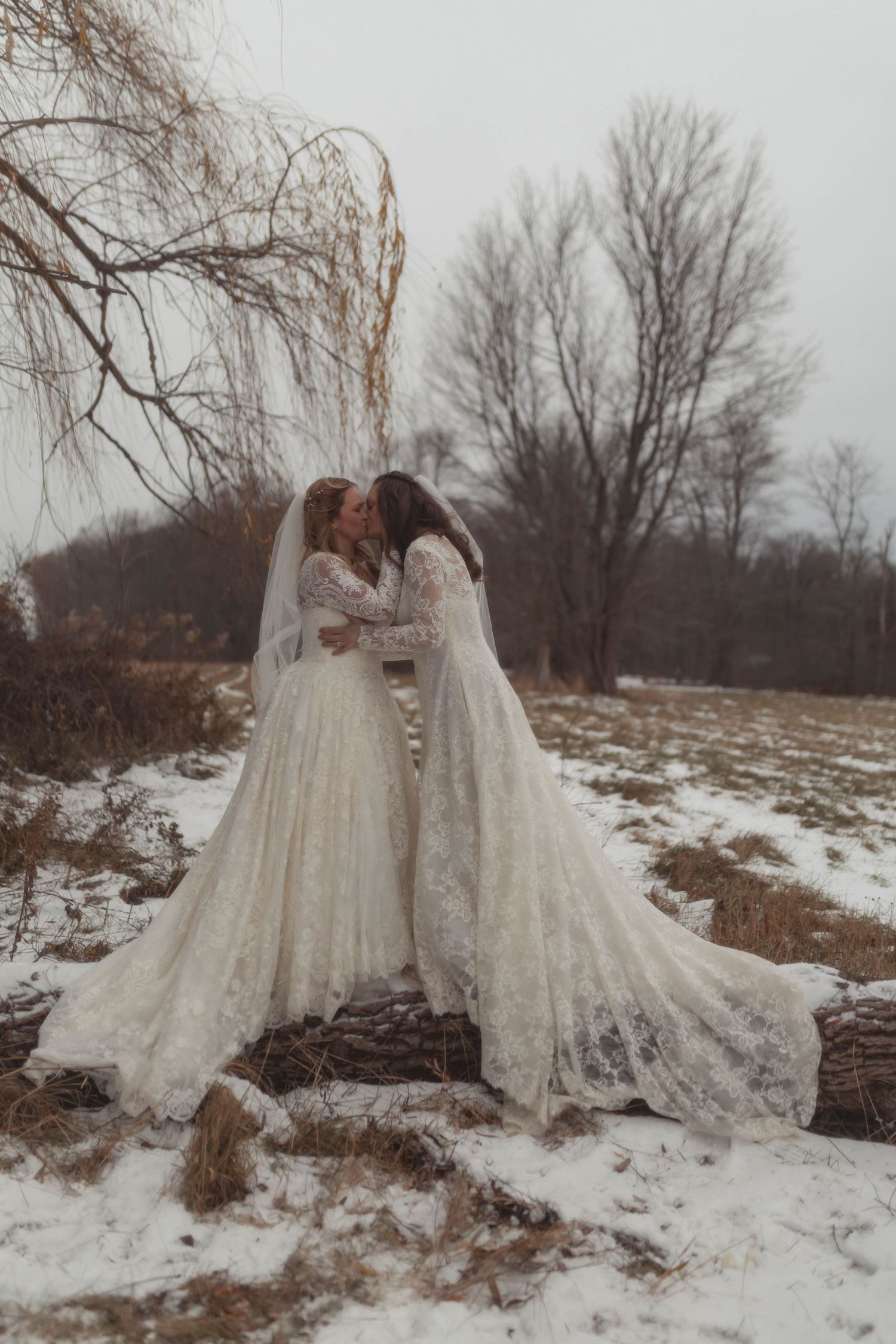 Two brides in lace wedding gowns share a kiss in a snowy winter landscape with bare trees.