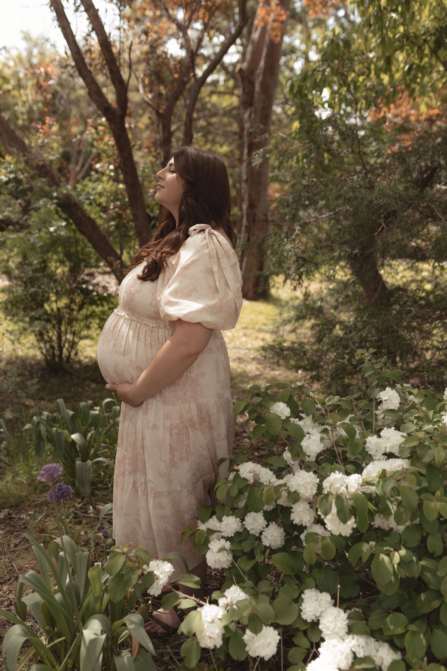 Pregnant woman in floral dress standing among white flowers and lush greenery in a serene garden setting.