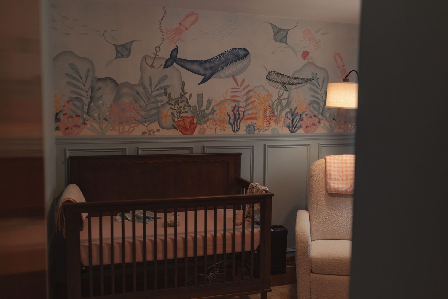 Serene nursery with wooden crib, ocean mural featuring whales, warm lamp light, and cozy glider chair.