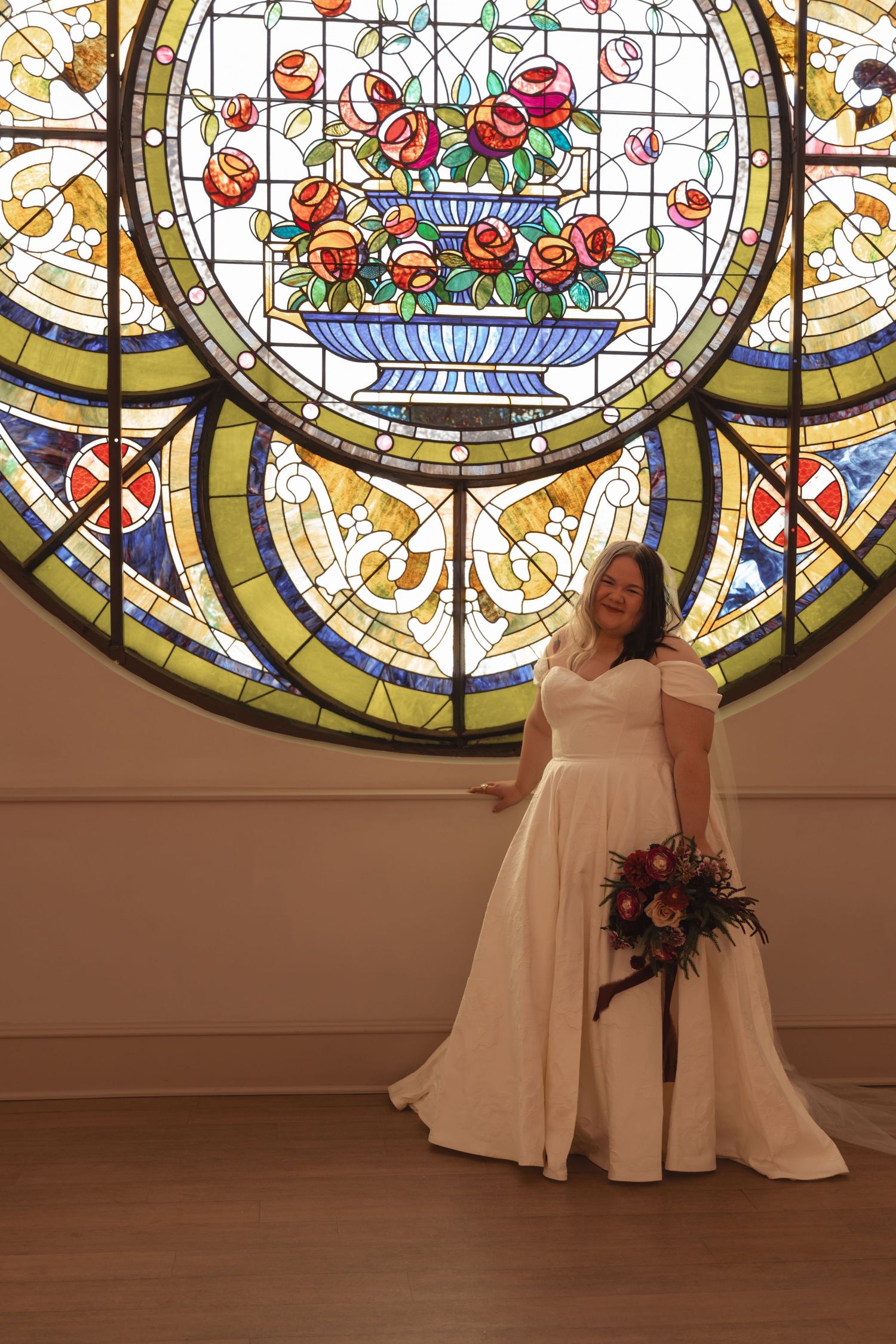 Bride in white off-shoulder gown holding bouquet before colorful stained glass window with floral motif.