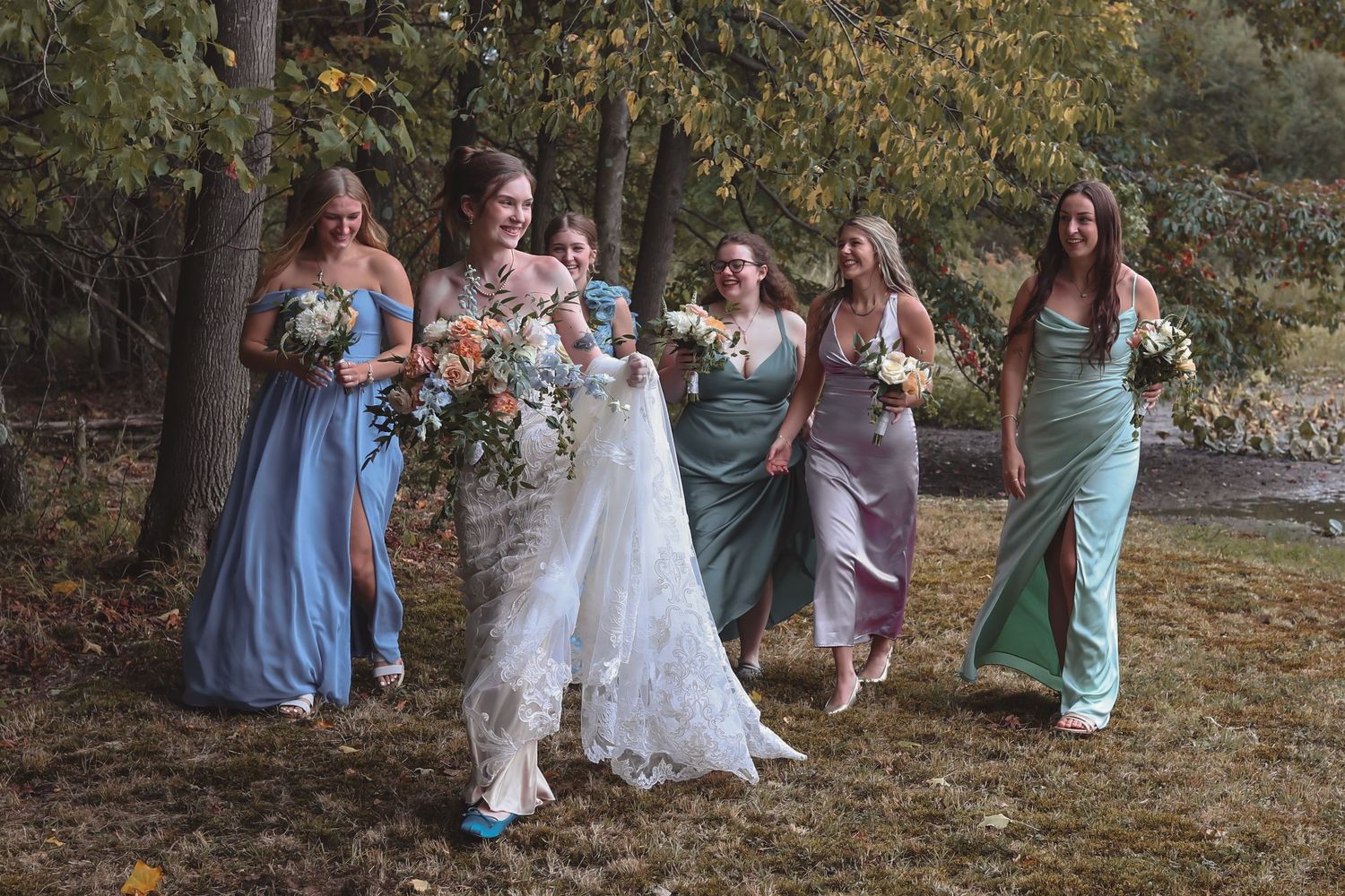 Bride in white lace gown walks with six bridesmaids in colorful mismatched dresses, holding bouquets outdoors.