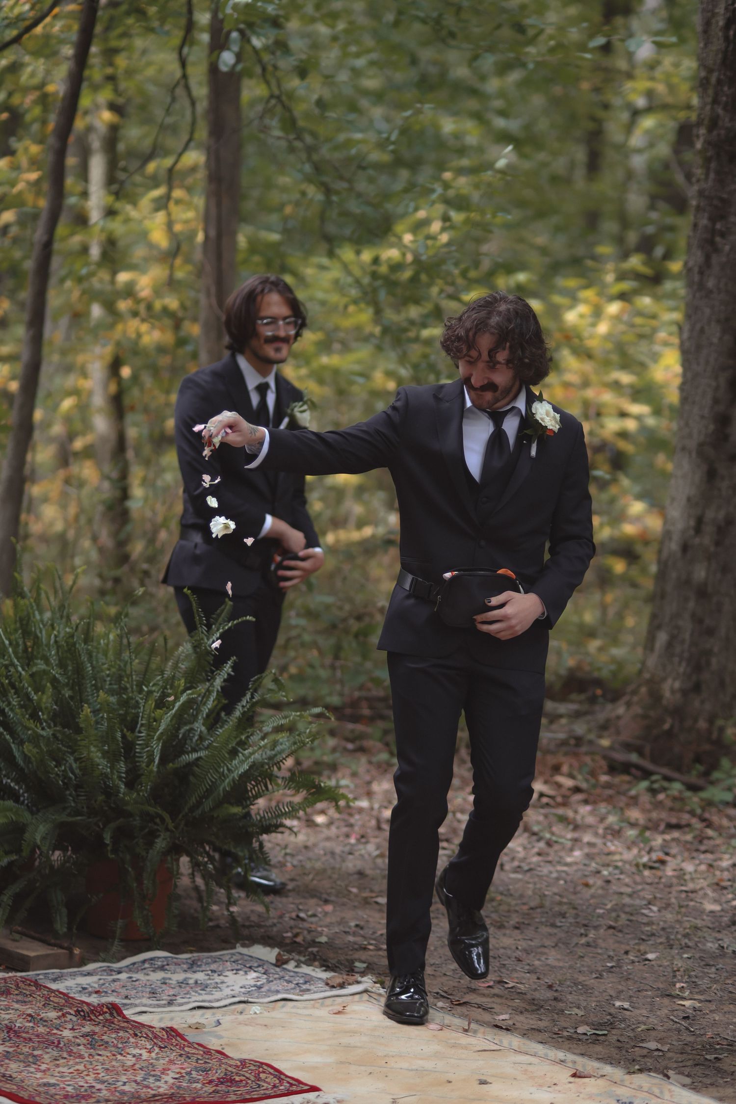 Two grooms in black suits walk down forest aisle scattering flower petals at outdoor wedding ceremony.