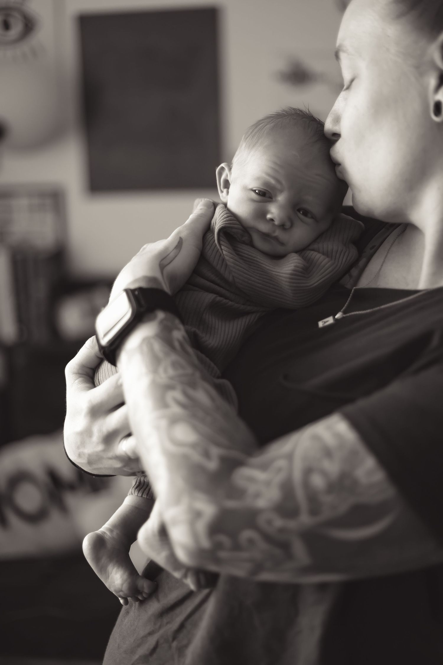 A tender black and white portrait of a parent with visible tattoos holding a sleepy infant in their arms.