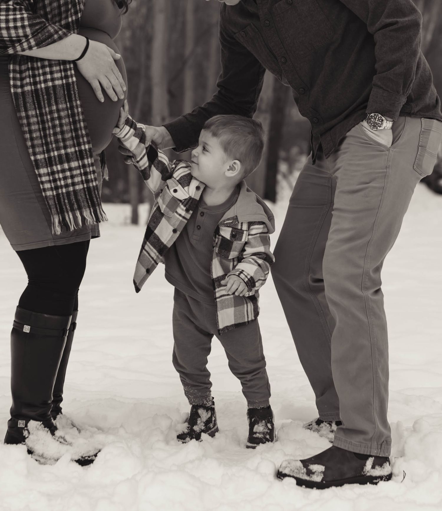 Black and white photo of a toddler in plaid jacket holding parents hands while standing in snow.