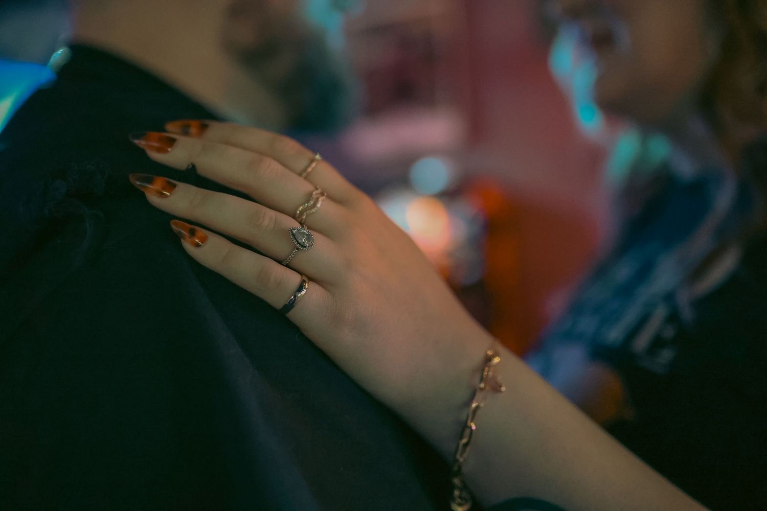 Close-up of a woman's hand with red nails, a diamond ring, and a gold bracelet resting on a dark jacket.